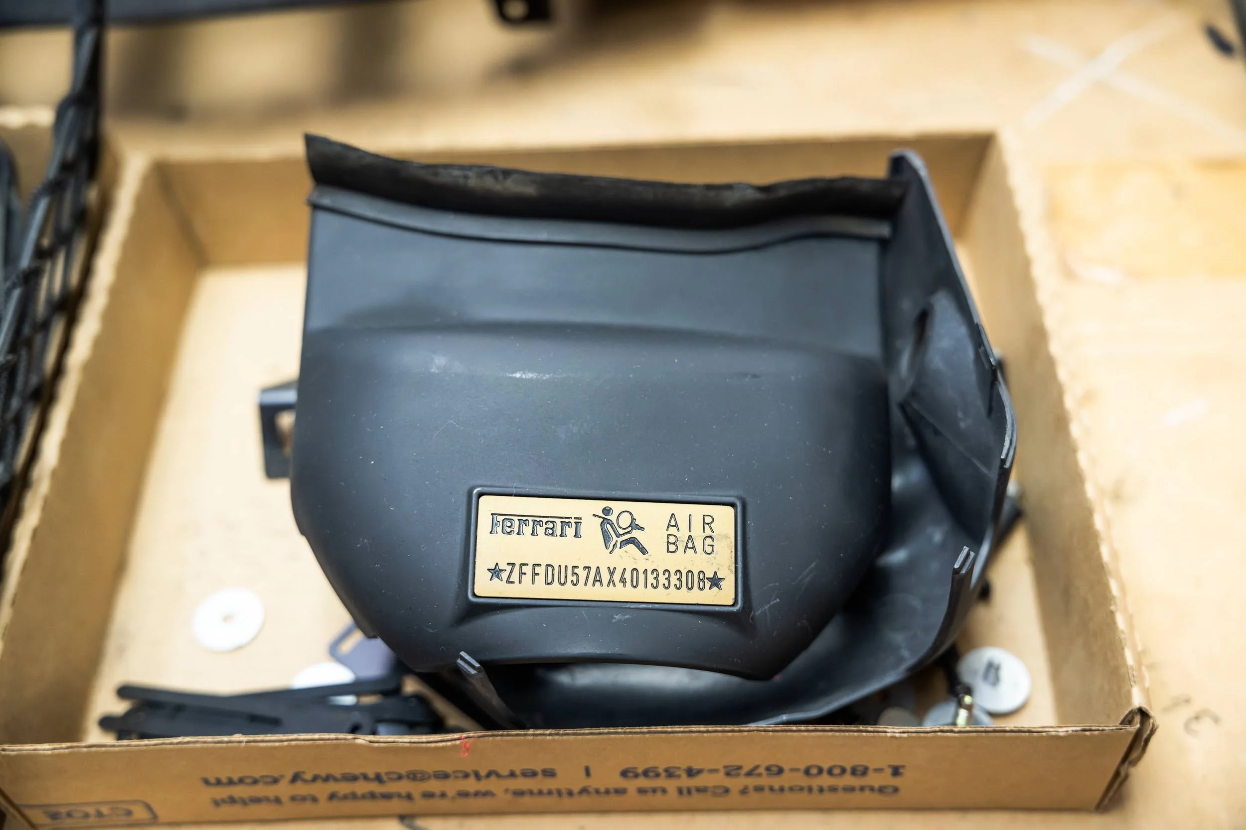 A black Ferrari airbag with a metal label showing the vehicle's serial number, placed inside a cardboard box.