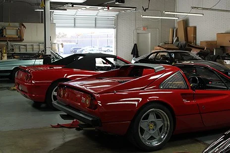 Two red vintage sports cars parked inside a garage, with one car's rear facing the camera and the other facing sideways.
