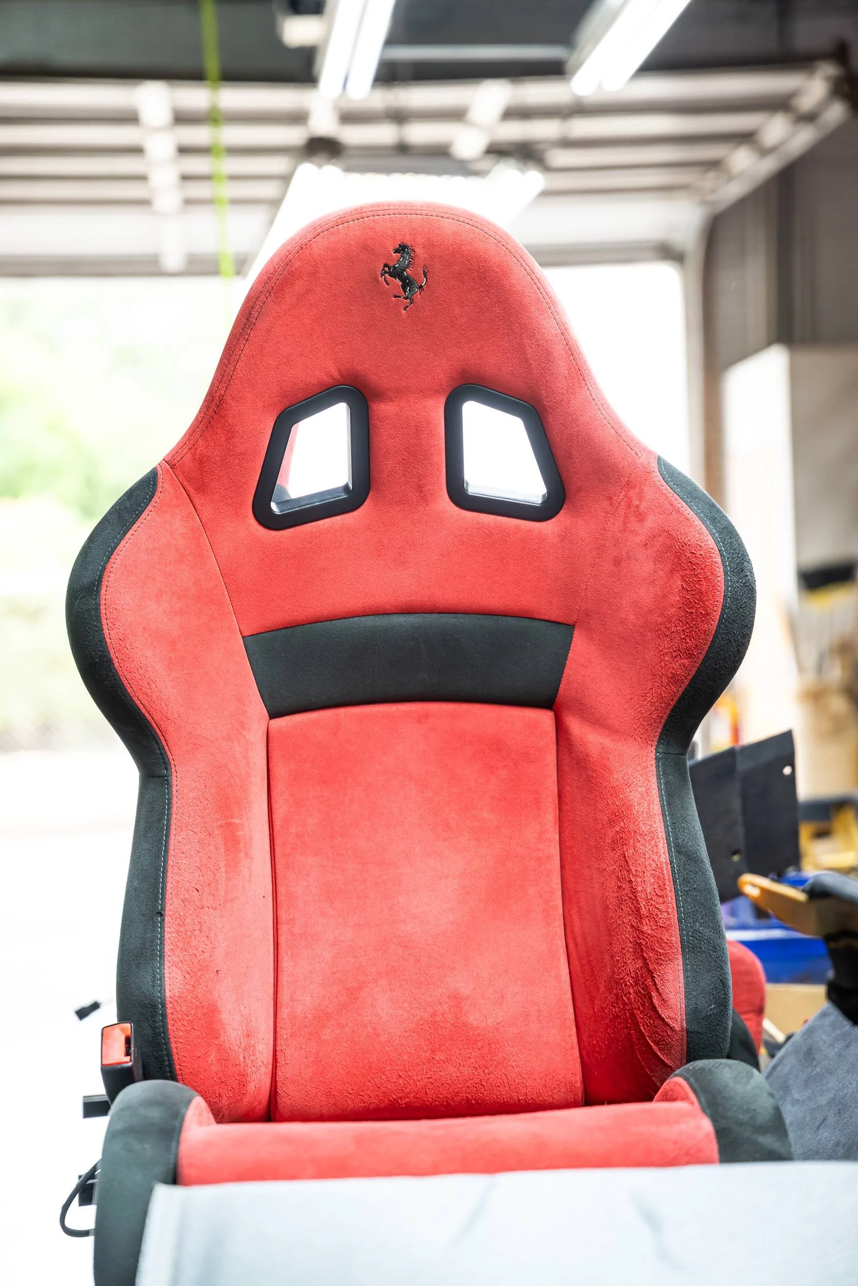A red and black racing car seat with a prancing horse logo on the headrest, in a workshop or garage setting.