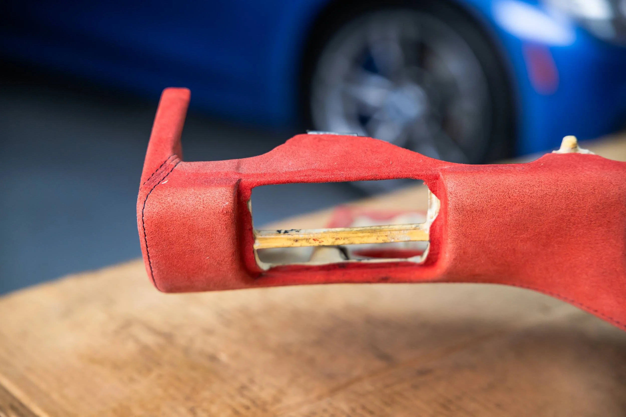 Close-up of a red leather object with stitching detail and a rectangular opening, placed on a wooden surface with a blurred blue and black background.