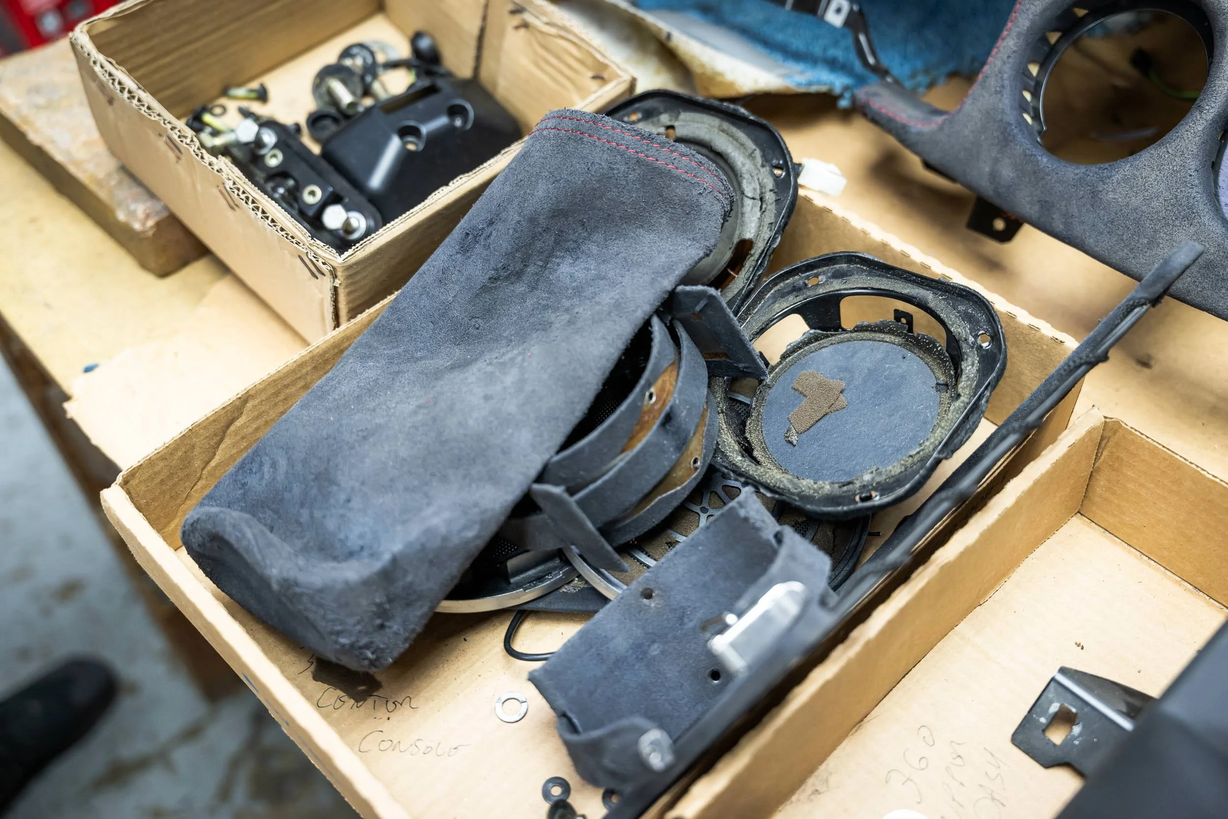 A cardboard box filled with disassembled and worn-out car parts, including a pair of goggles, metal brackets, and plastic pieces, on a workbench.