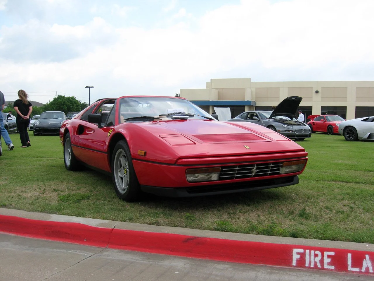 A red vintage Ferrari sports car parked on a grassy area at a car show, with several other exotic cars visible in the background.