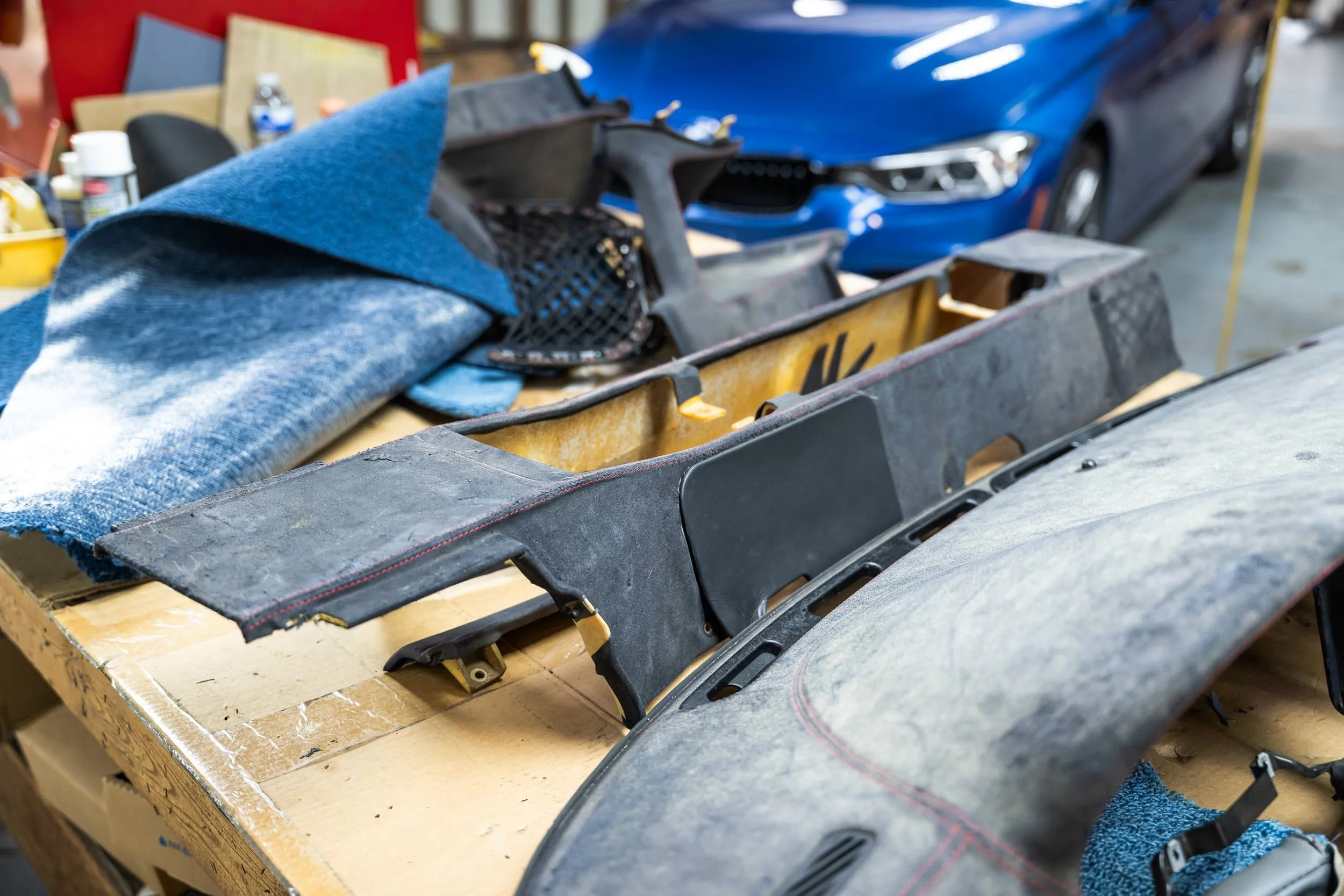 A workbench with car interior parts, a blue towel, and a blue sports car in the background at an auto repair shop.