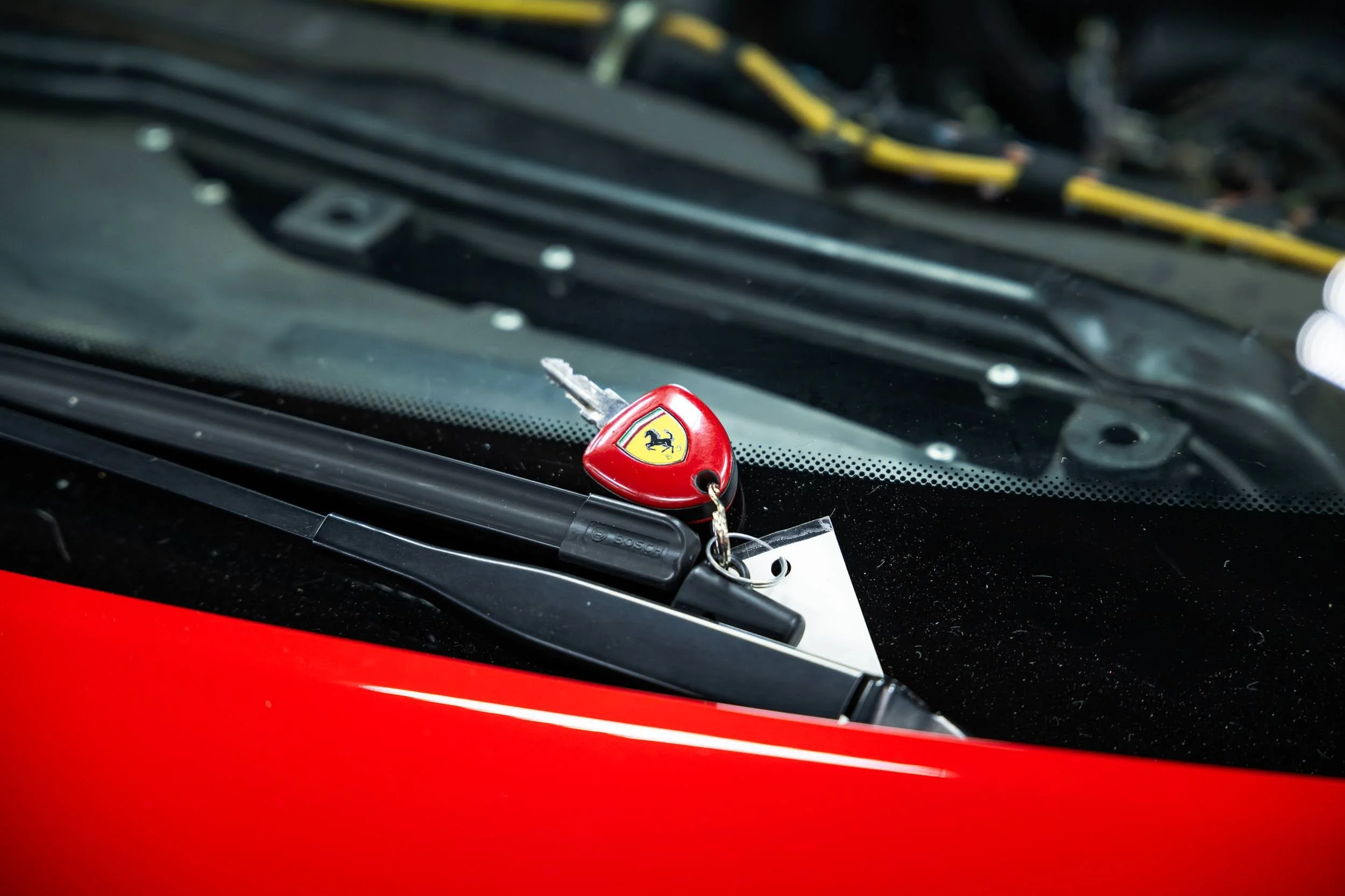 Close-up of a red Ferrari keychain with the Ferrari logo hanging on a car dashboard, with the windshield and some wires visible inside the vehicle.