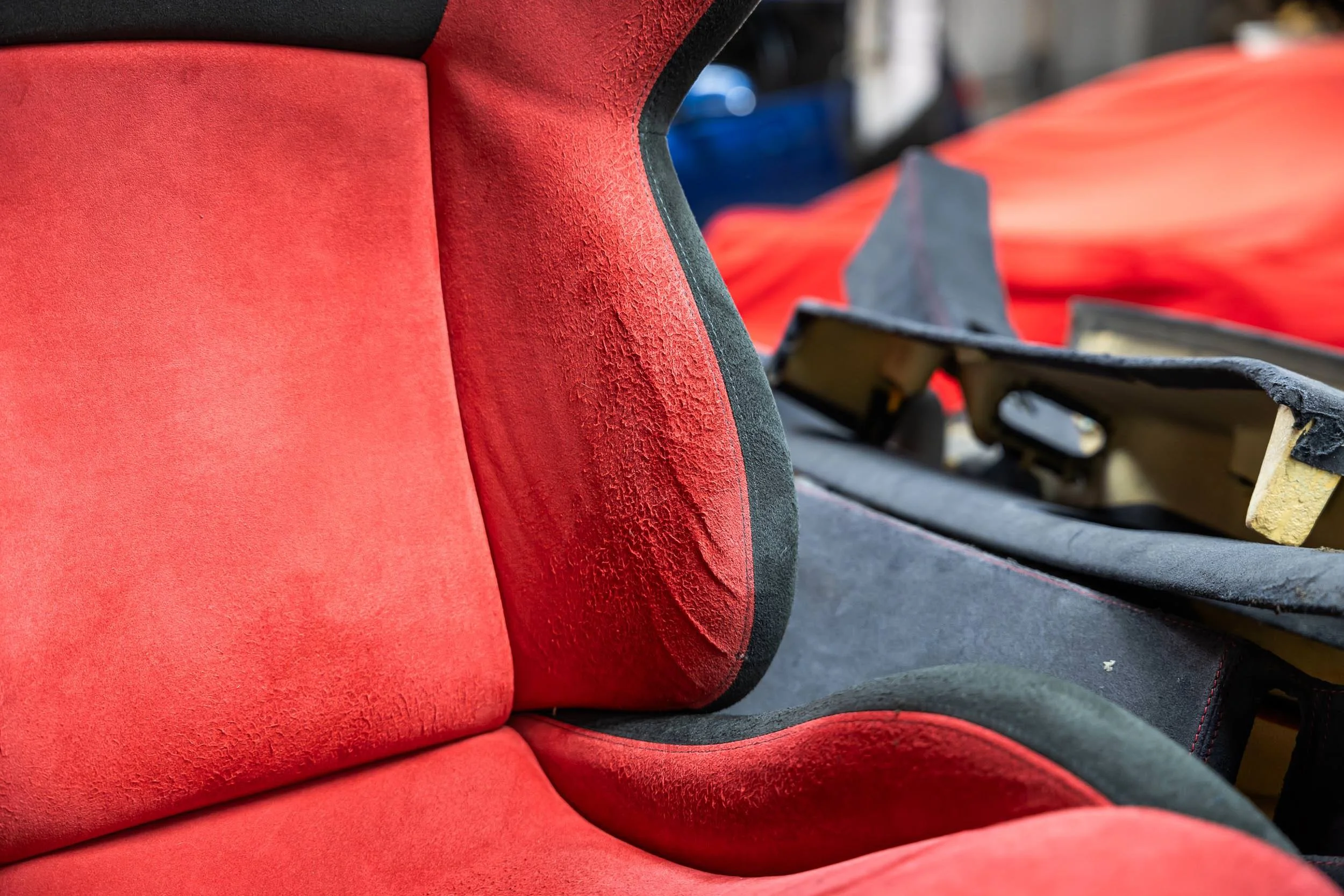 Close-up of a red sports car seat with visible texture and black accents, with part of a black and yellow component and some blurred background elements.
