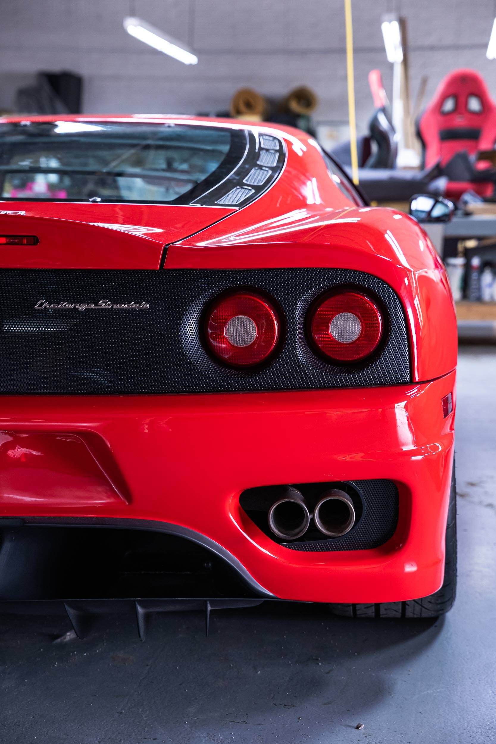 Close-up of the rear end of a red sports car, showcasing the taillights, rear bumper, and dual exhaust pipes, inside a garage with tools and equipment visible in the background.
