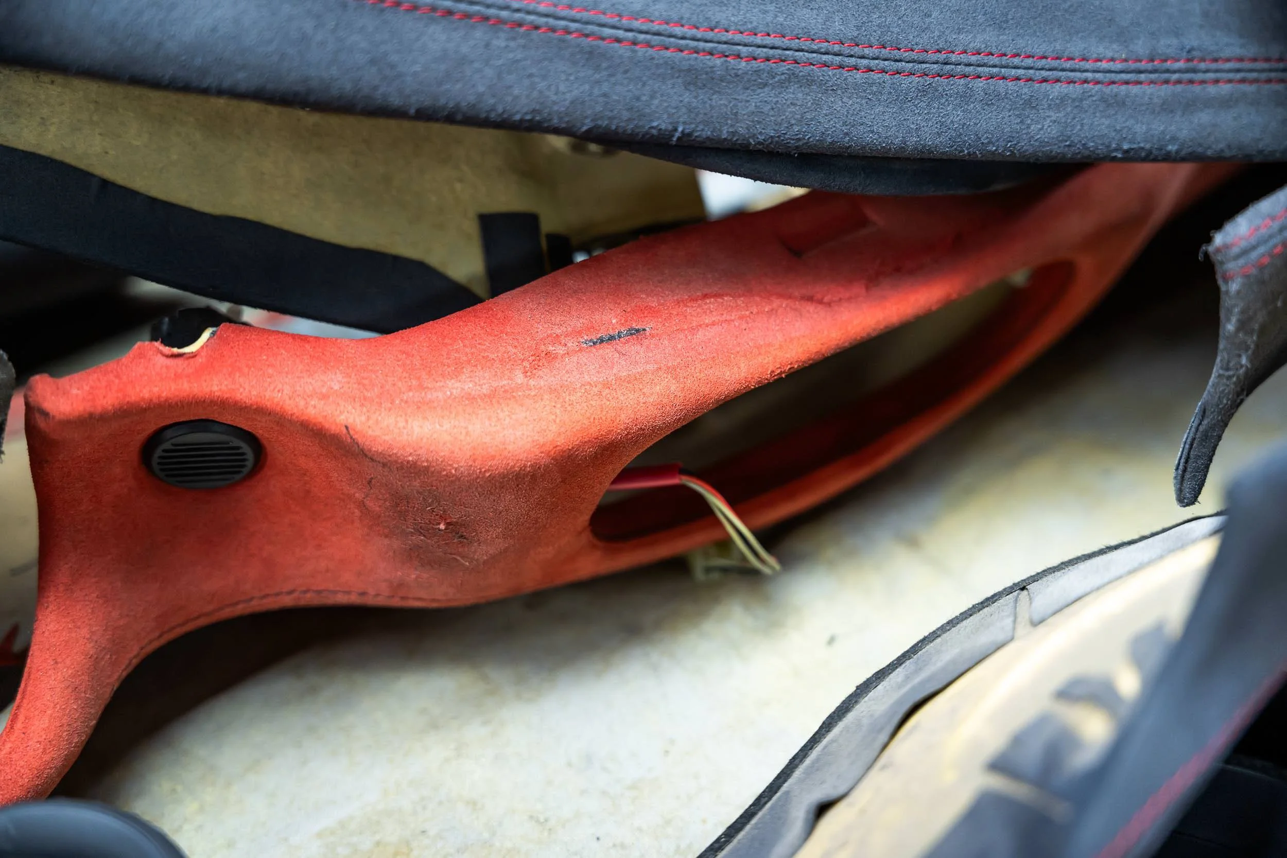 Close-up of the underside of a seat, showing a red metal bracket, a small black speaker, and wires, with portions of gray and black upholstery and a yellowish foam padding visible.