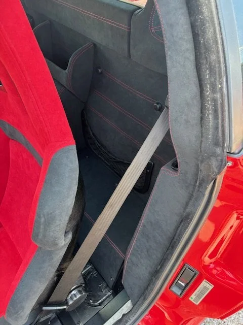 The back seat of a red car with a gray fabric interior, showing a beige seat belt hanging across the center area, with the right side of the car's interior visible.