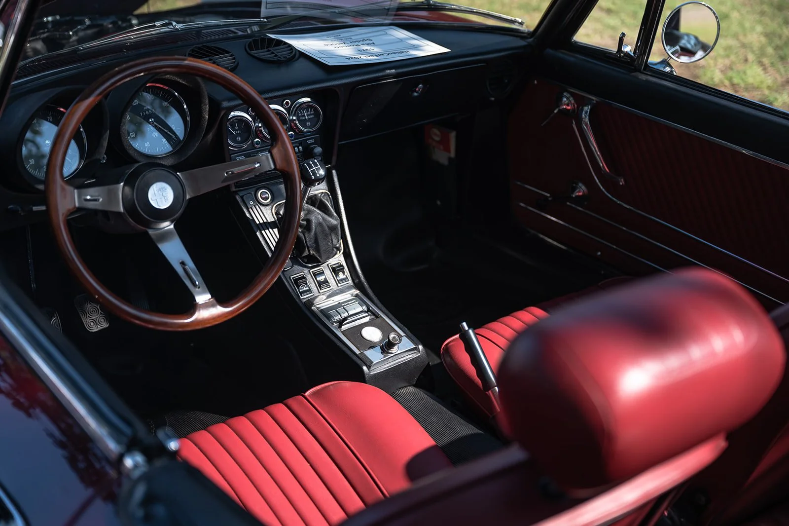 Interior of a classic car with a wooden steering wheel, red leather seats, a manual gear shift, and a dashboard with multiple gauges and controls.