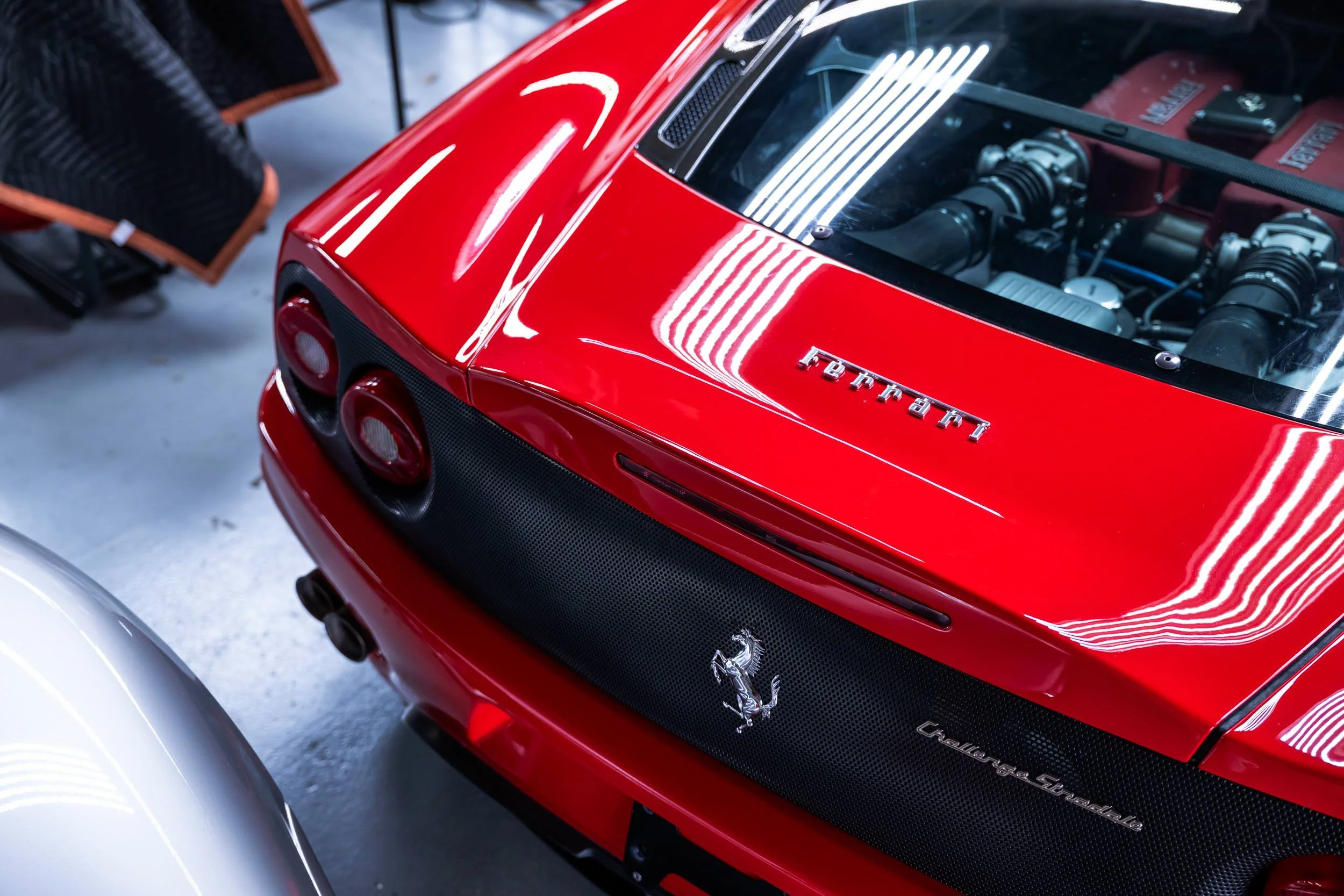 A close-up of the rear of a red Ferrari sports car, showing the Ferrari logo, engine details through a transparent cover, and part of the rear grille and tail lights in a garage or showroom.