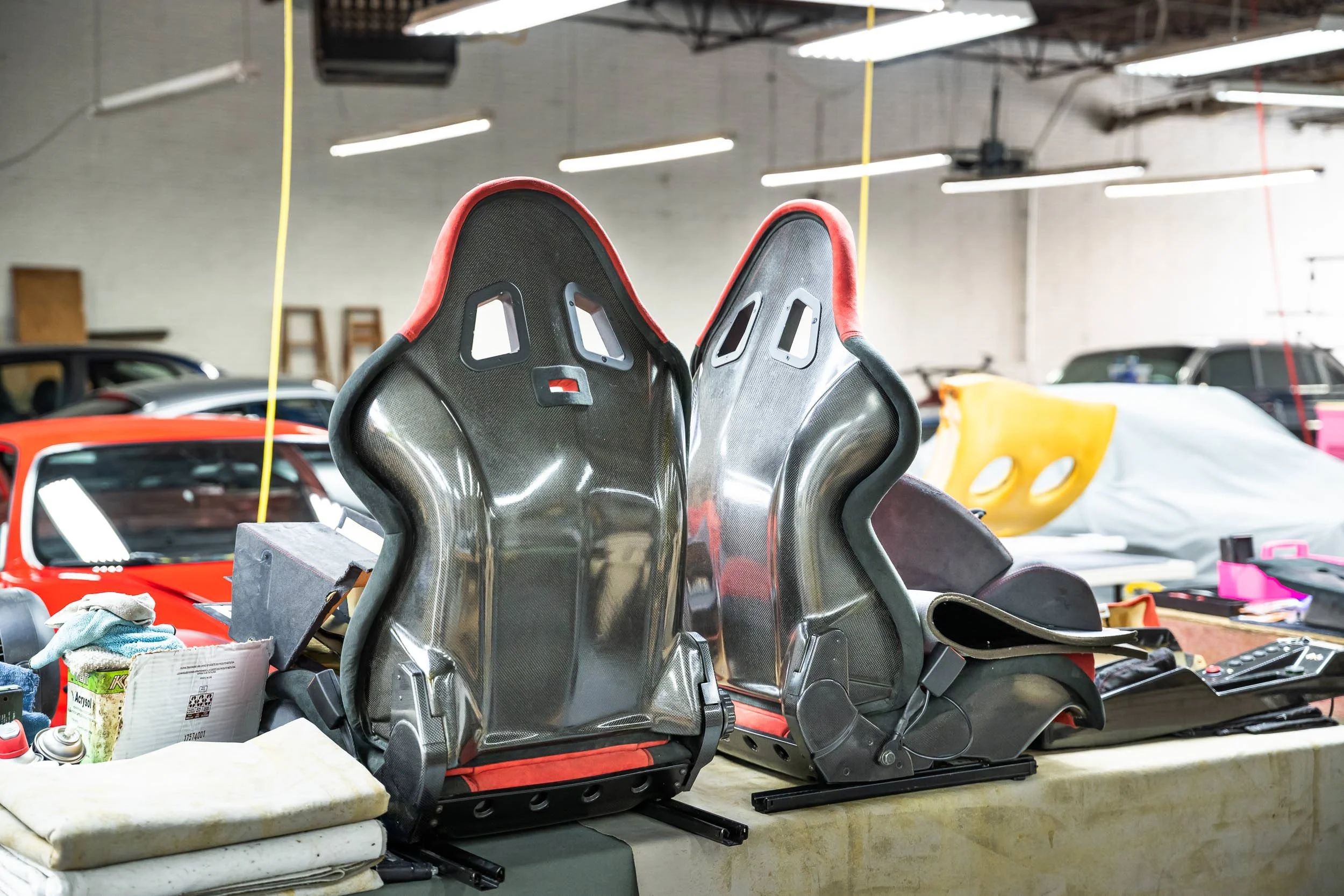 Several carbon fiber racing seats with red trim placed on a workbench in a garage or workshop surrounded by various tools, car parts, and supplies.