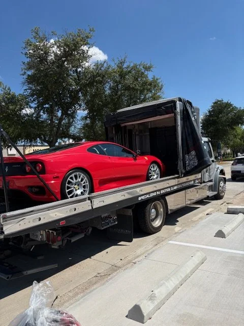 A red sports car is loaded onto a flatbed tow truck parked on a street with trees in the background.