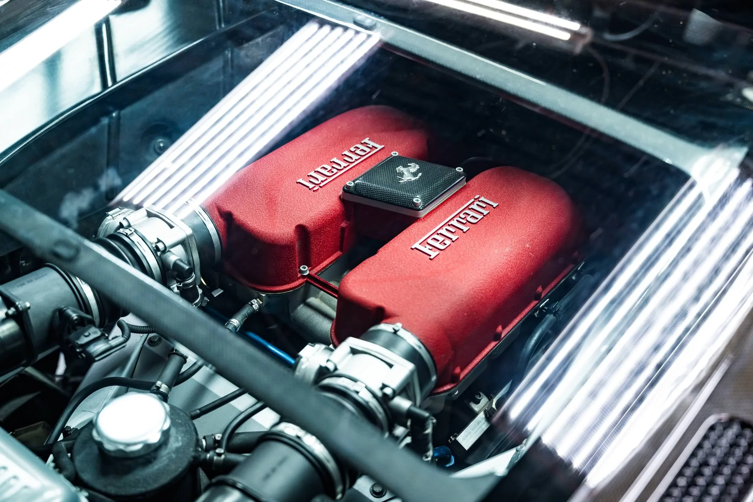 Close-up view of a Ferrari engine with red intake manifolds and carbon fiber components inside a car's engine bay.