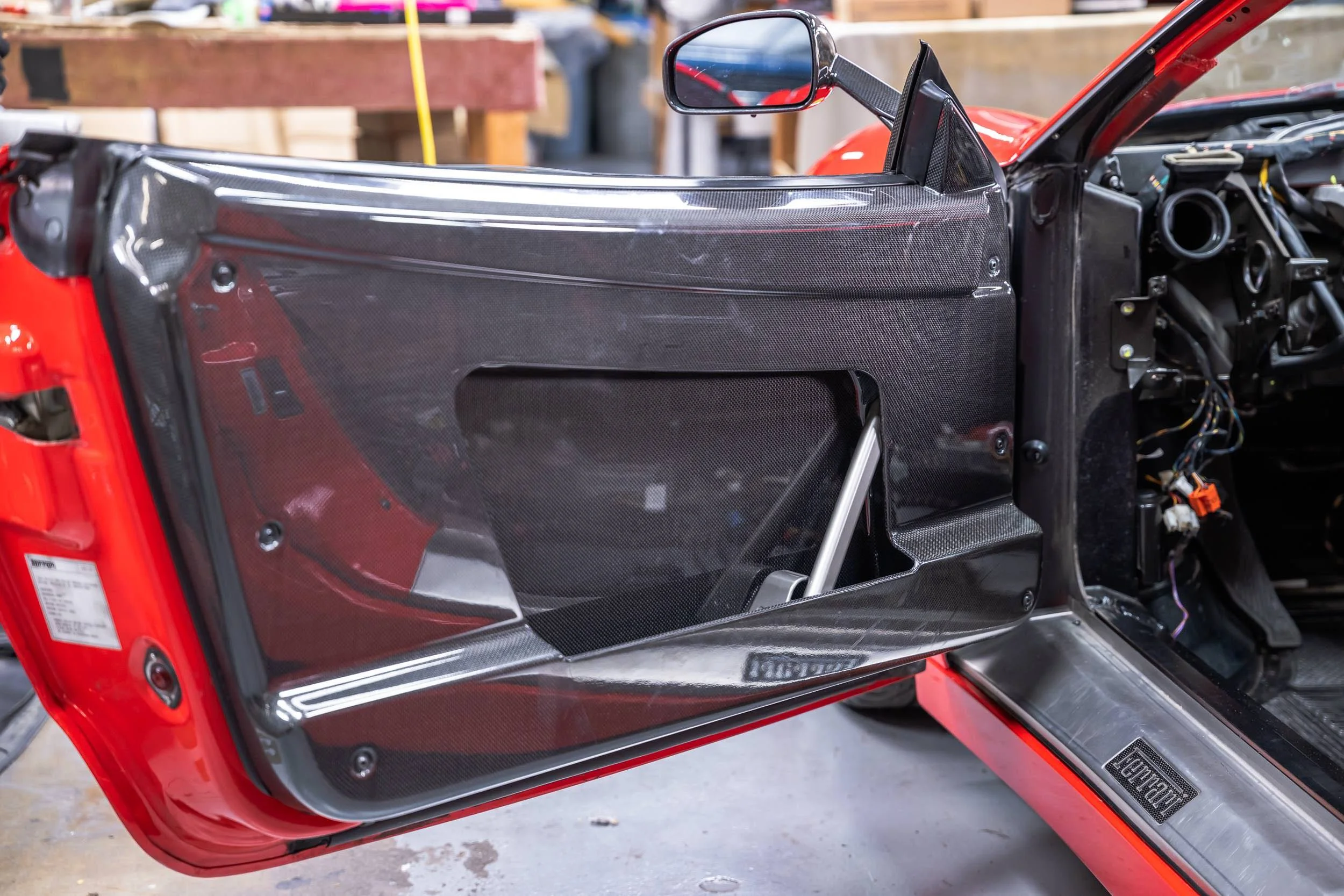 Close-up of a red race car with its driver's door open, revealing a carbon fiber interior and a side mirror.