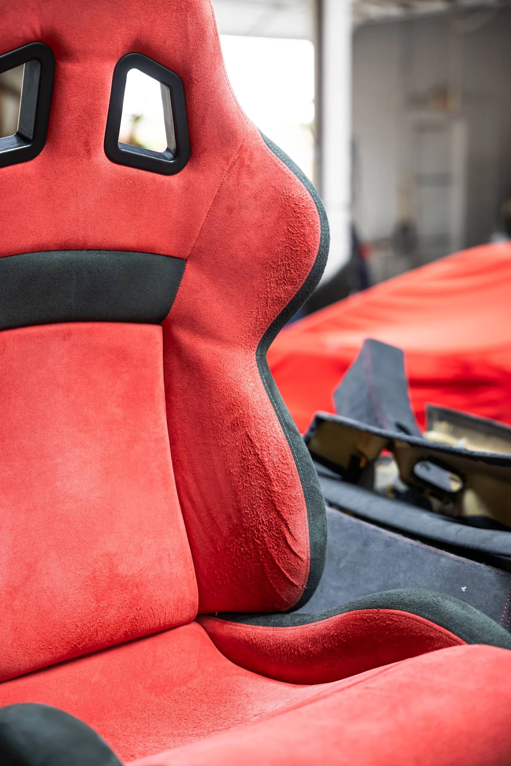Close-up of a red racing seat with black harness openings, showing textured and smooth fabric, in an automotive or racing setting.