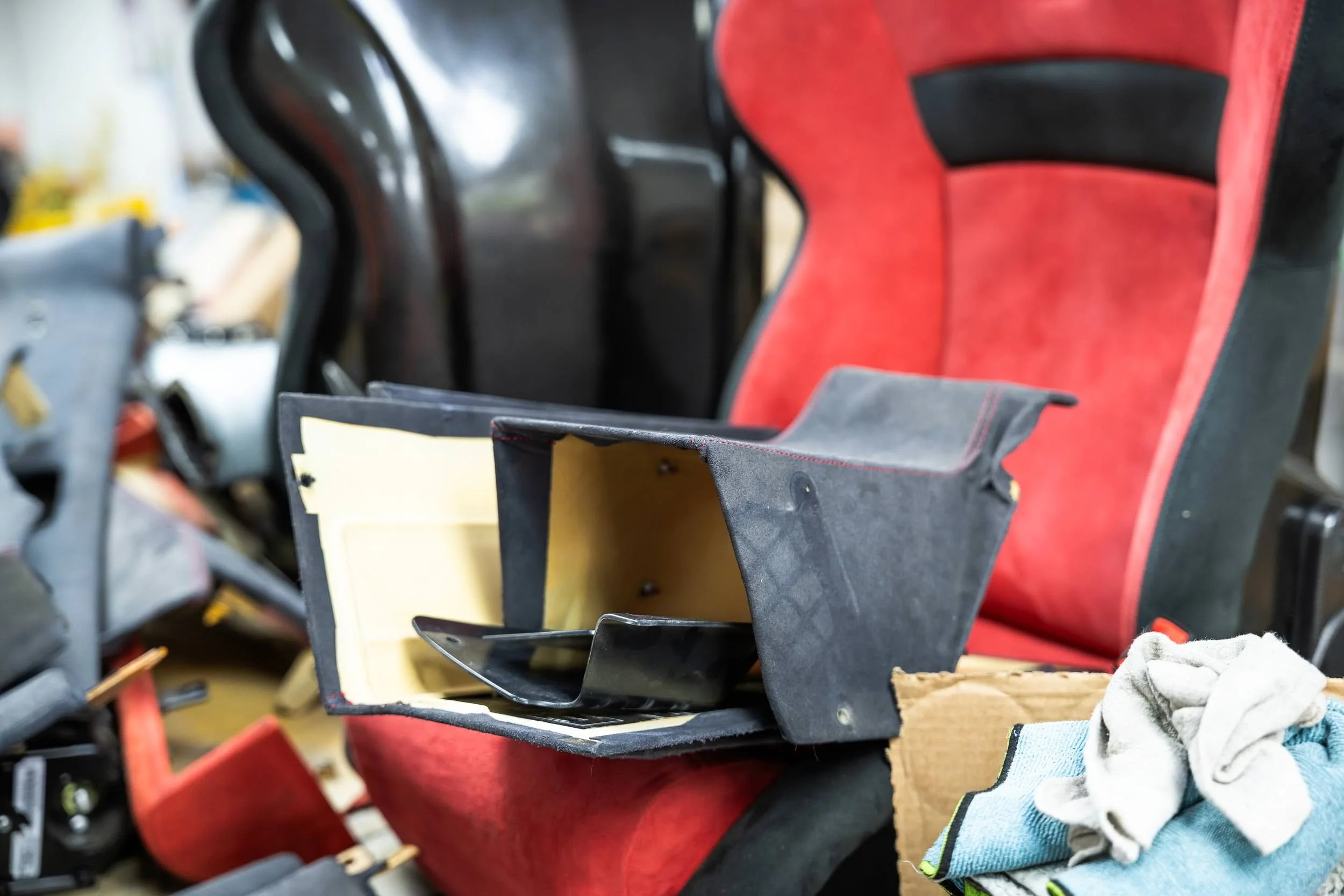 Close-up of a partially assembled black and yellow plastic object placed on a cluttered workbench with various tools, fabrics, and a red chair in the background.
