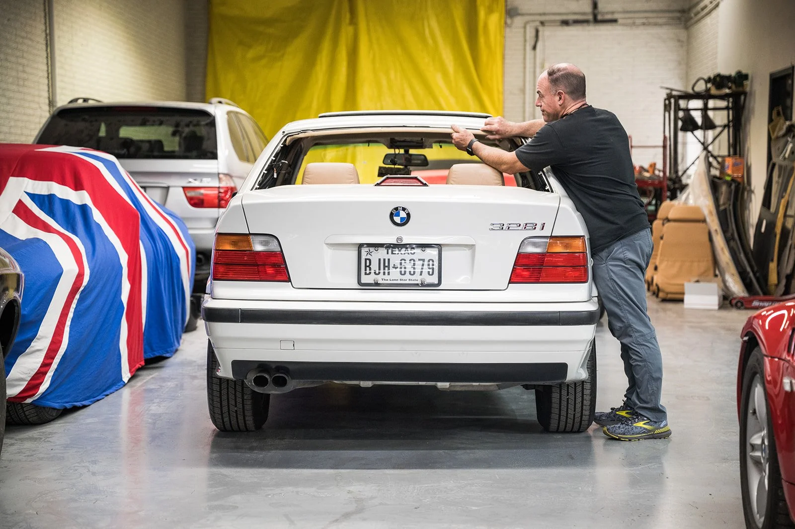 A man working on a white BMW 3 Series car in an indoor garage, with two other cars covered and several tools in the background.