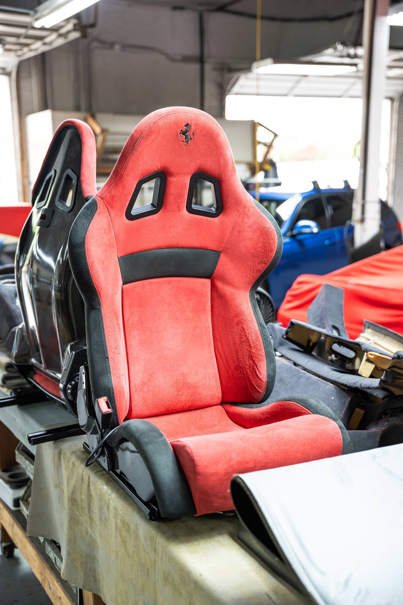 A red racing car seat with black accents and a Ferrari logo on the headrest, placed on a worktable in a garage or workshop with other car parts and vehicles in the background.