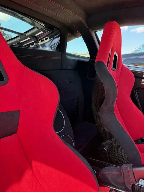 Interior of a sports car showing two red racing bucket seats with black accents, a portion of the dashboard, and a clear blue sky visible through the windows.