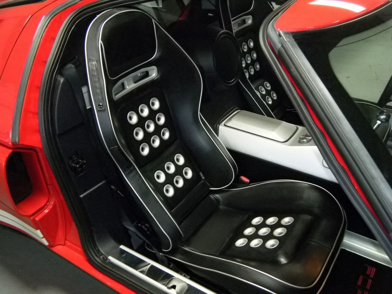 The interior of a vintage red sports car with black seats that have white circular holes and white piping, a white center console, and a black dashboard.