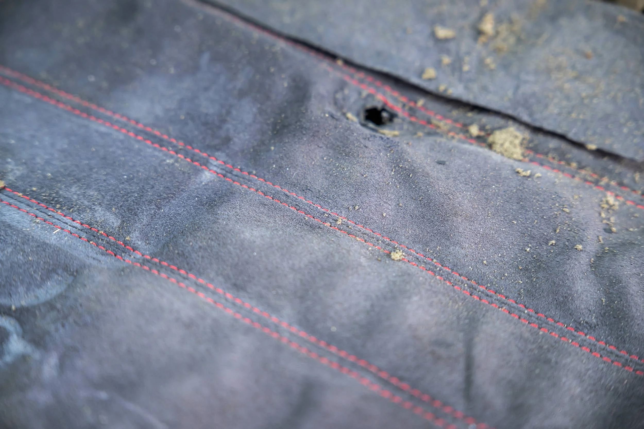 Close-up of black fabric with red stitching and a small, round hole in the material, with dirt and debris around the hole.