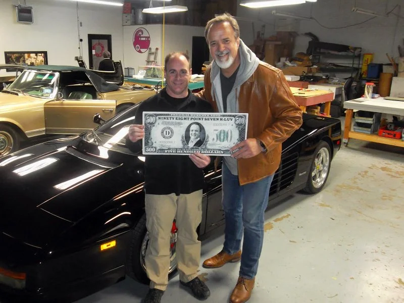 Two men standing in a garage, holding a large novelty dollar bill, next to a black sports car.