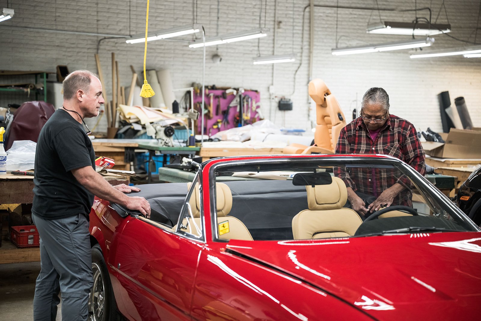 Two men working on a red convertible car in a workshop. The workshop has tools, a workbench, a mannequin, and various supplies in the background.