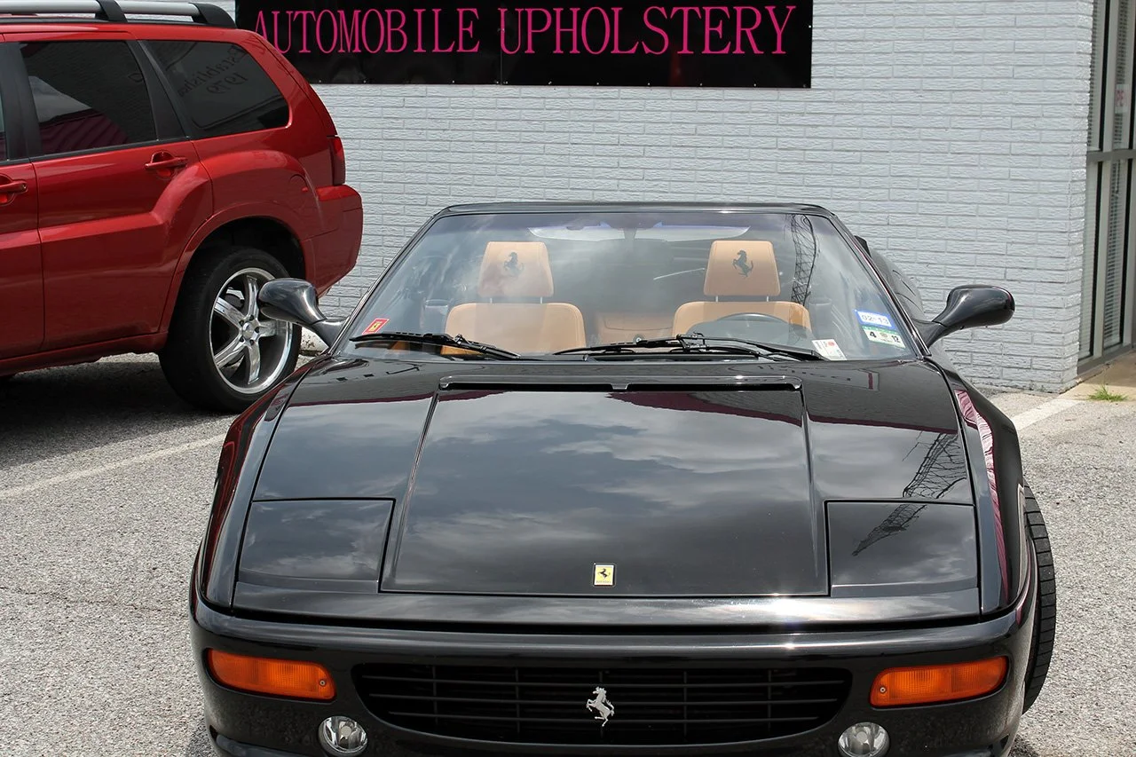 A black Ferrari convertible parked in front of a gray brick wall with a sign that reads 'Automobile Upholstery.' The car has tan interior seats and a transparent windshield, with a red vehicle parked next to it.