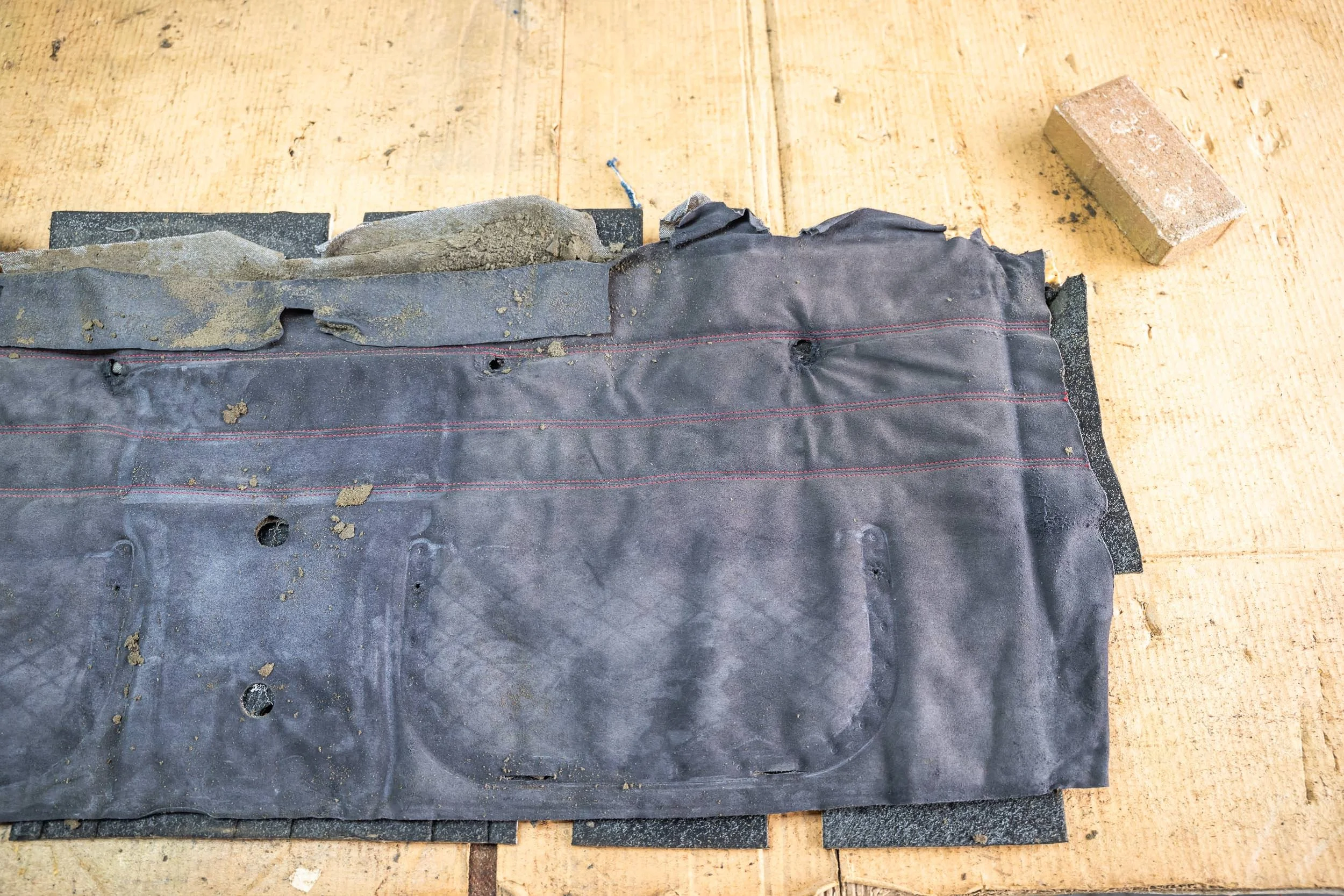 Close-up of a damaged black rubber sheet or mat with holes and tears, secured to a wooden surface with a brick nearby.