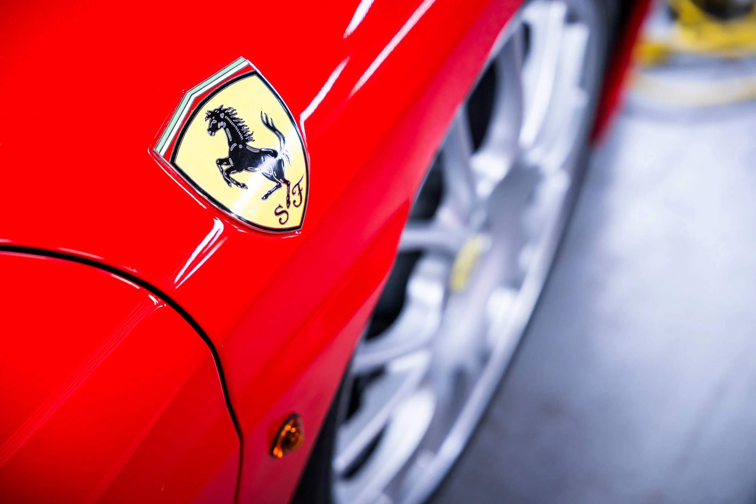 Close-up of a red Ferrari car showing the Ferrari emblem logo with a prancing horse on a yellow background, with part of the car's wheel in the background.
