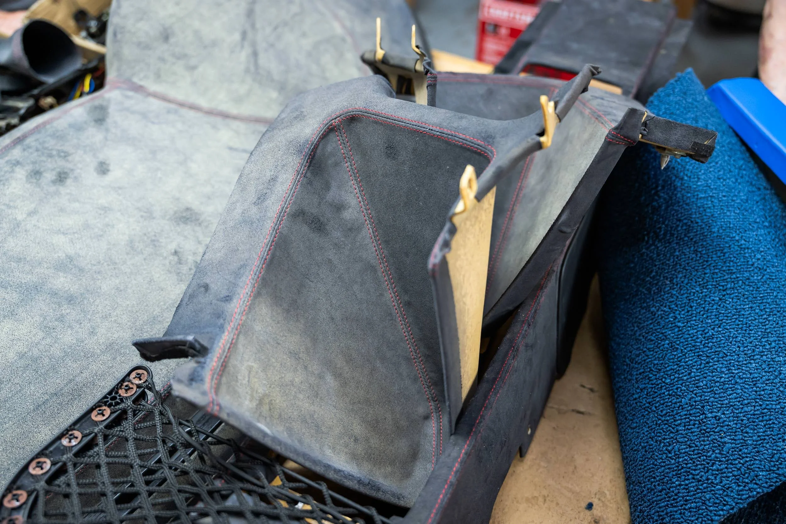 Close-up image of automotive fabric upholstery in a workshop setting, showing black and grey fabrics with red stitching, secured with golden clips, with a blue towel and a black plastic grid in the foreground.