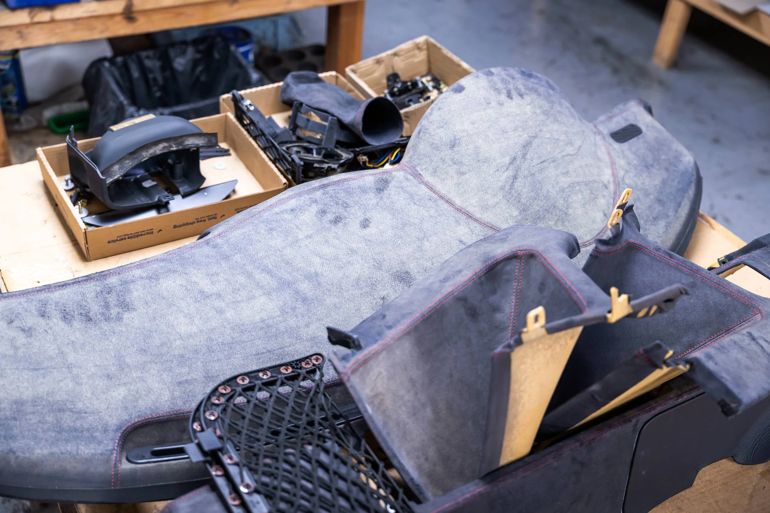 A workbench with a padded automotive seat, a black plastic component, a cardboard box with various black parts and wires, and a black plastic grille with multiple circular openings.