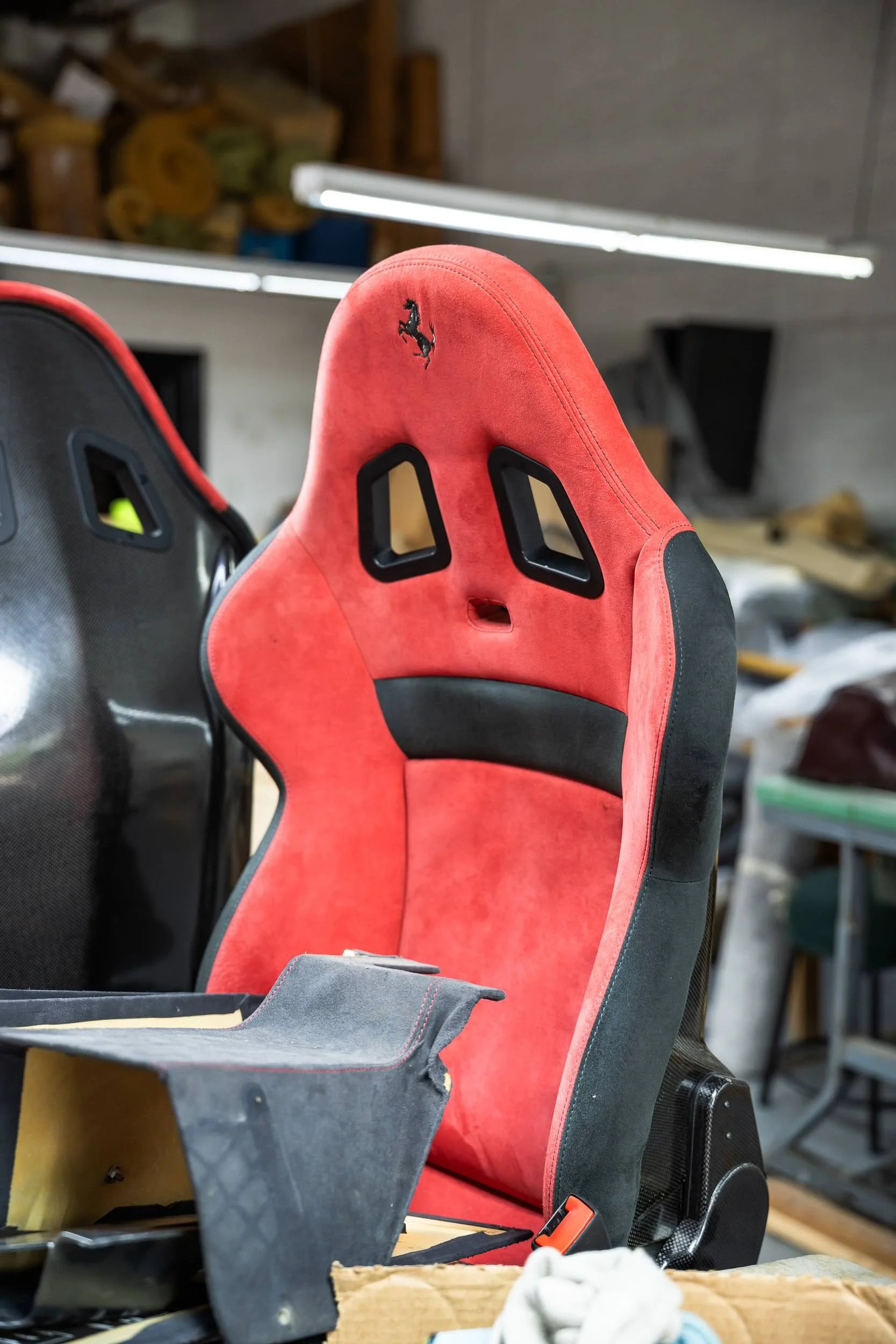 A red racing car seat with black accents and a prancing horse logo on the headrest, situated in a workshop or garage environment with wooden shelves and fluorescent lighting.