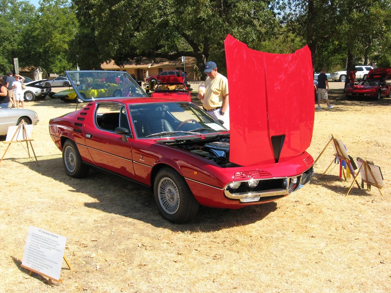 Red classic sports car with an open hood, displayed at an outdoor car show, surrounded by other vintage cars and spectators.