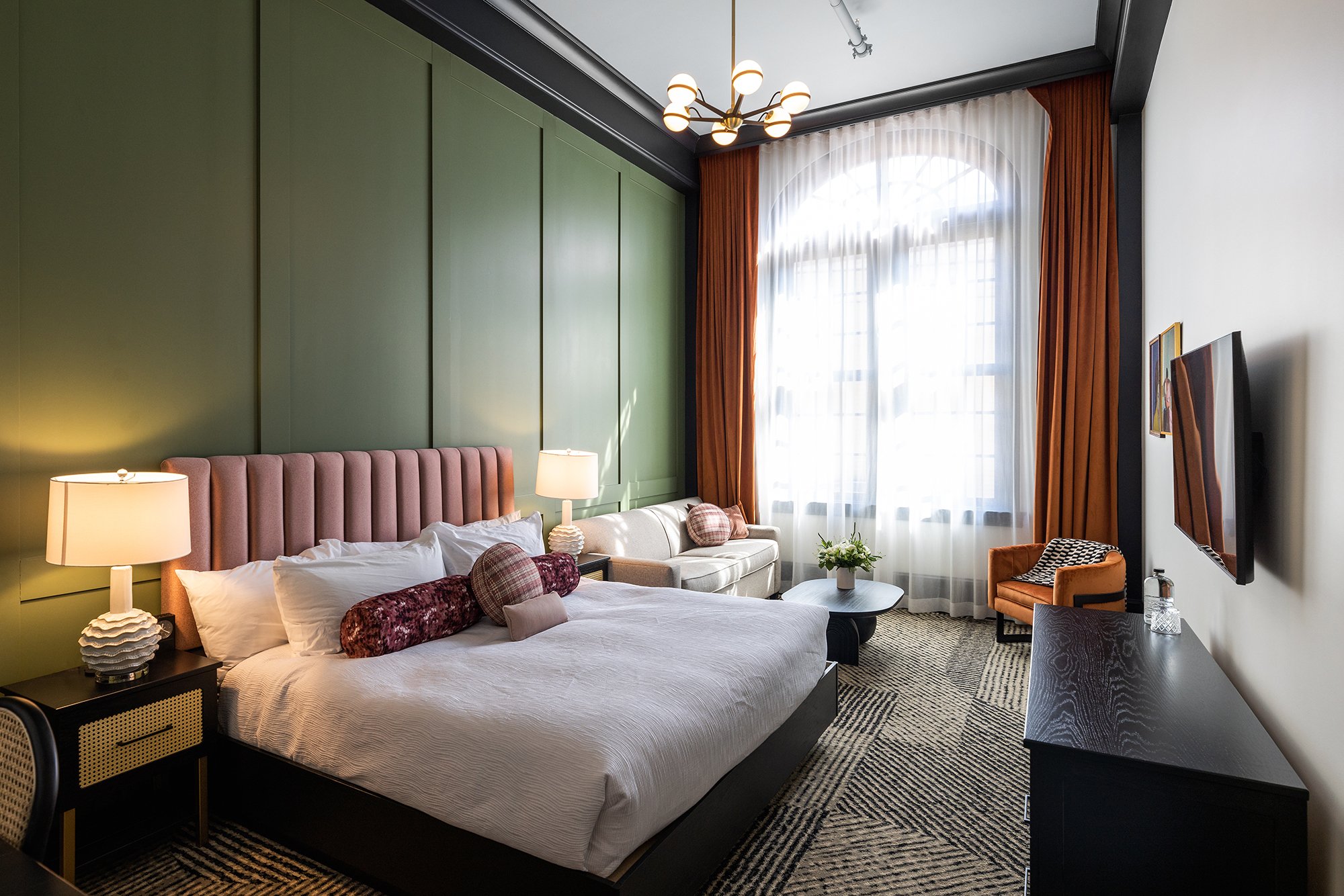 Hotel room with a large bed, green accent wall, pink headboard, two bedside lamps, a sofa with pillows, a round coffee table, a large window with sheer white curtains and orange drapes, a wall-mounted TV, and a patterned rug.