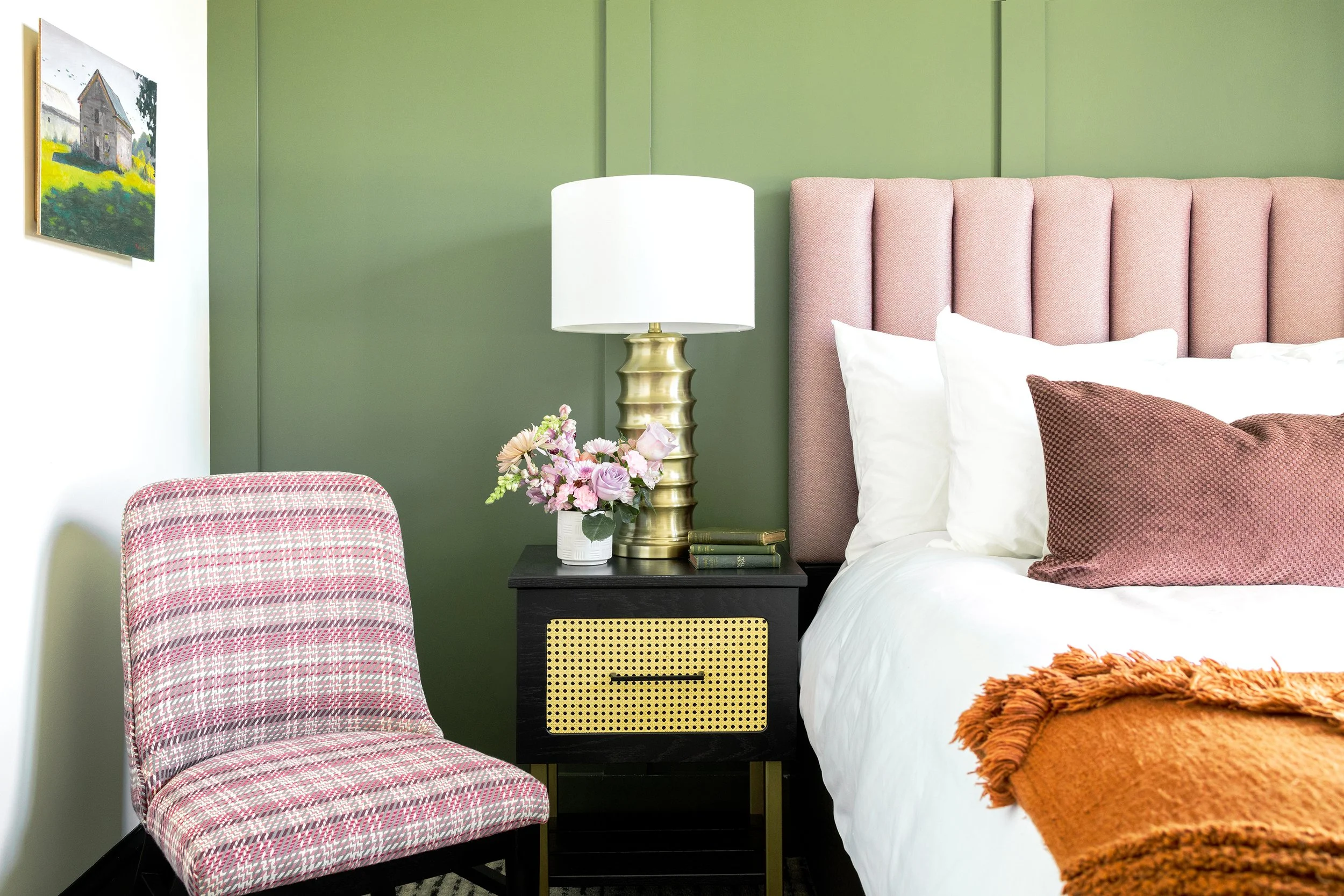 Bedroom with green wall, pink upholstered headboard, white bedding, decorative pillows, orange throw blanket, black nightstand with yellow woven drawer, white lamp with gold base, pink floral arrangement, pink patterned chair, and framed painting of 