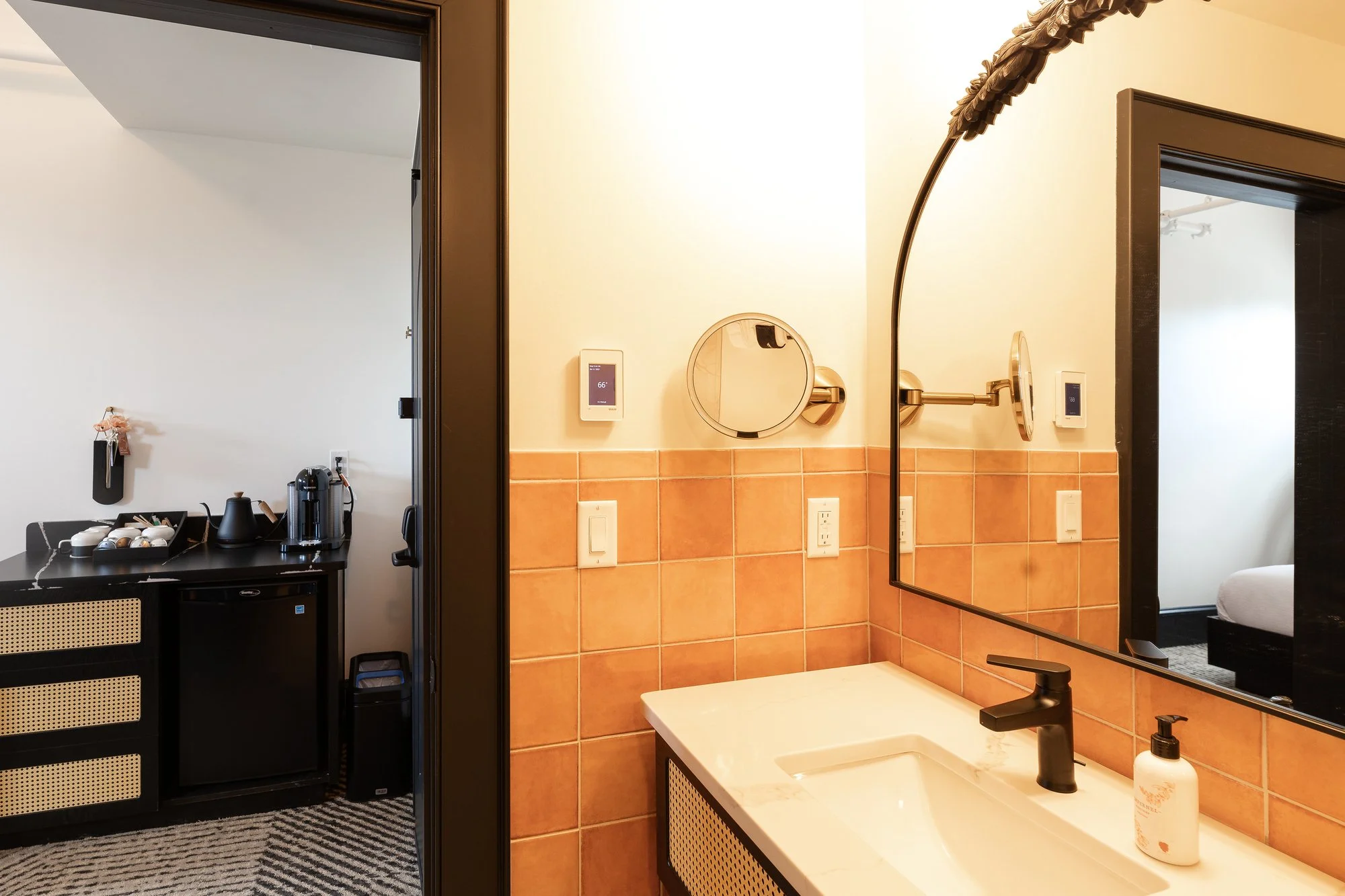 A bathroom with a square white sink, black faucet, soap dispenser, and mirror, with a beige tiled wall and a large mirror reflecting part of a bedroom. An adjacent room has a black counter with coffee and tea supplies, and a door leading to another r