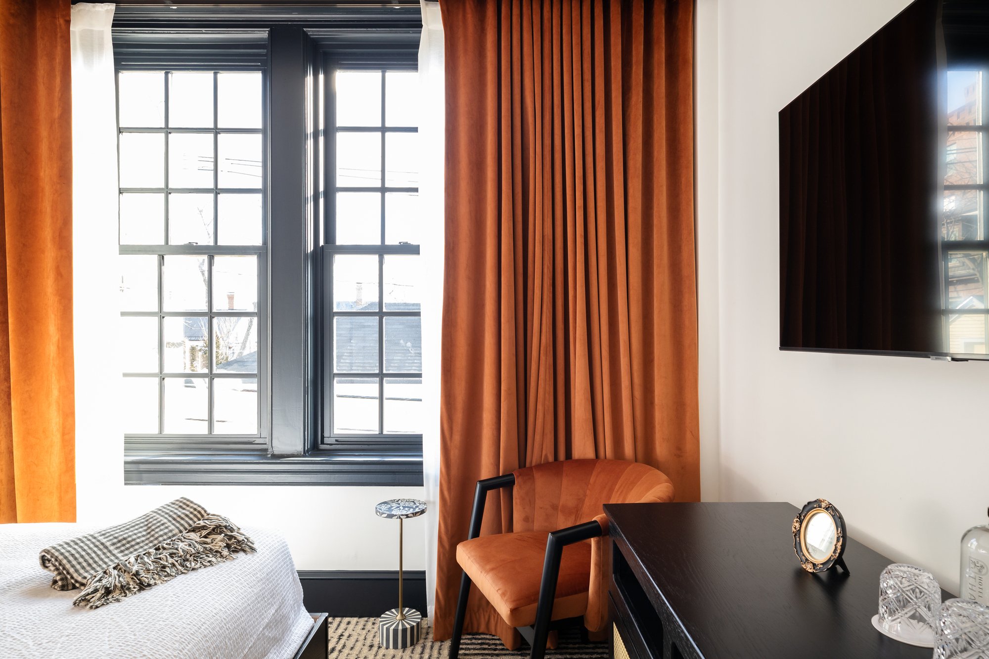 A cozy living room corner with a window framed by orange curtains, a velvet orange armchair, a small side table, a black dresser with decorative items, and a wall-mounted flat-screen TV.