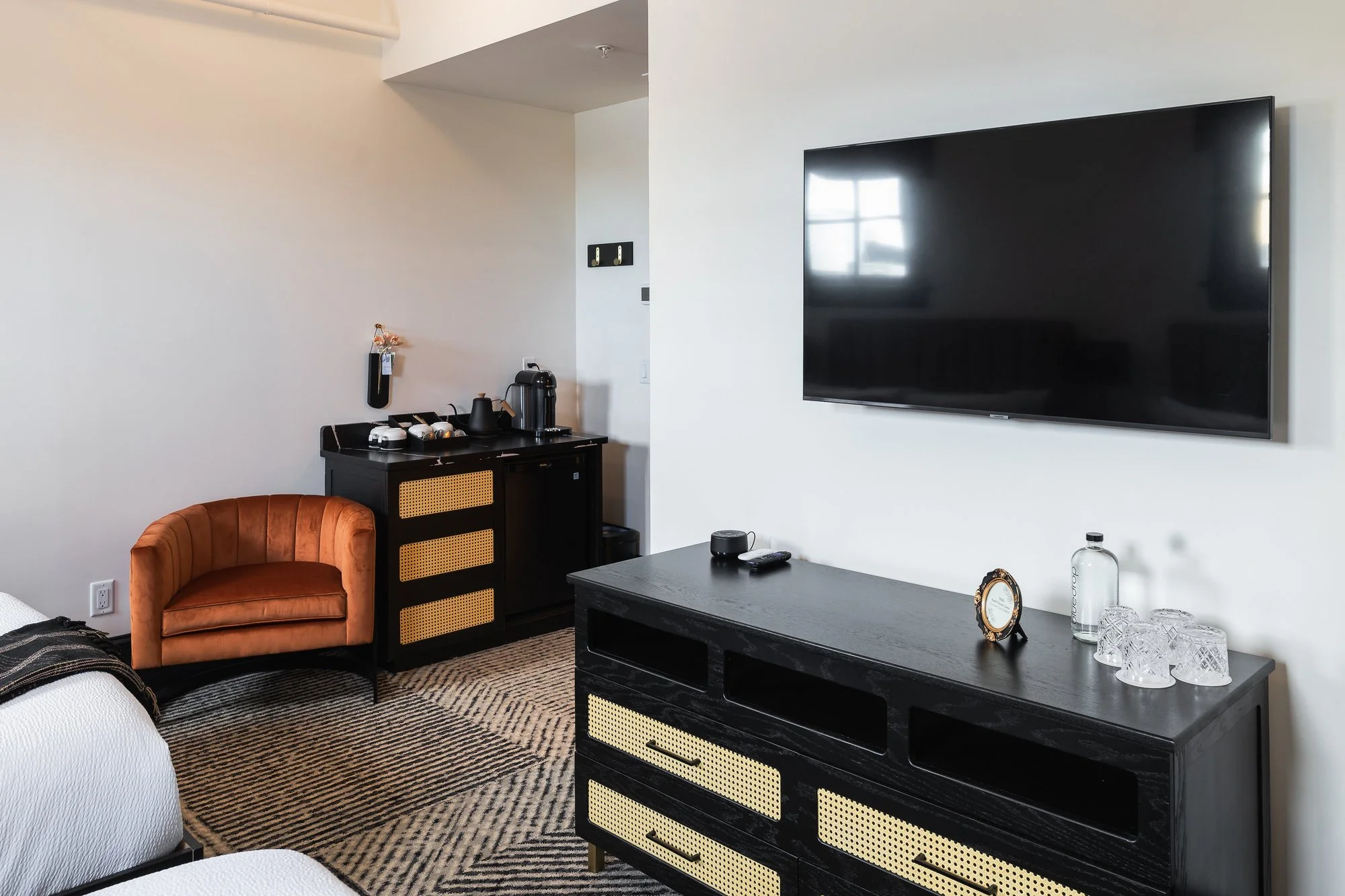 Hotel room with a black TV mounted on a white wall, a black cabinet with a remote and decorative items, a small orange velvet armchair, and a corner with a coffee station including a coffee maker, mugs, and water bottles, on a patterned rug.