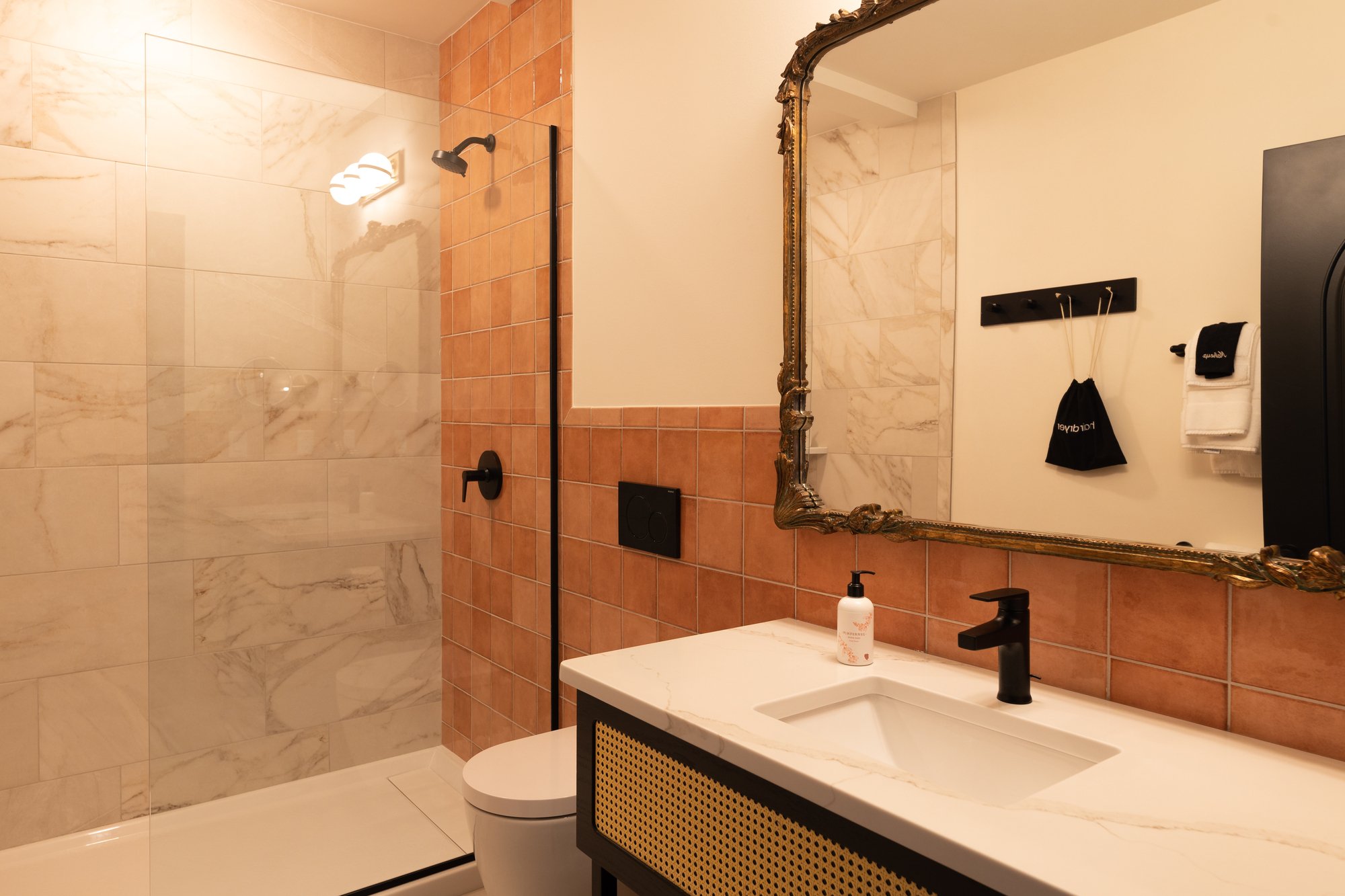 Modern bathroom with beige and terracotta tiles, glass shower with black fixtures, white toilet, and a white vanity with a marble countertop, gold-framed mirror, and a black faucet.