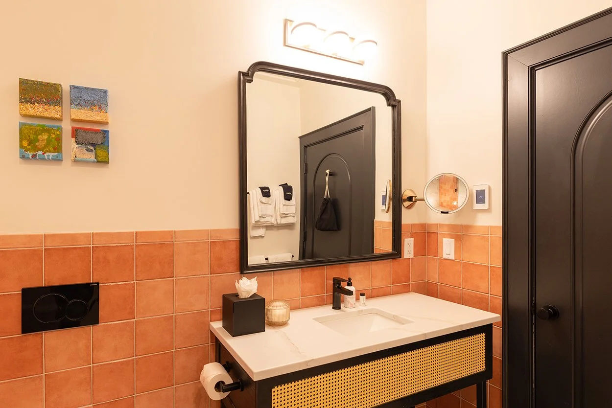 Bathroom with beige walls, orange tile halfway up, black-framed mirror above white sink, black light fixture, abstract paintings on wall, black cabinet with rattan front, towel hooks with towels, tissue box, candle, soap dispenser, small mirror on wa