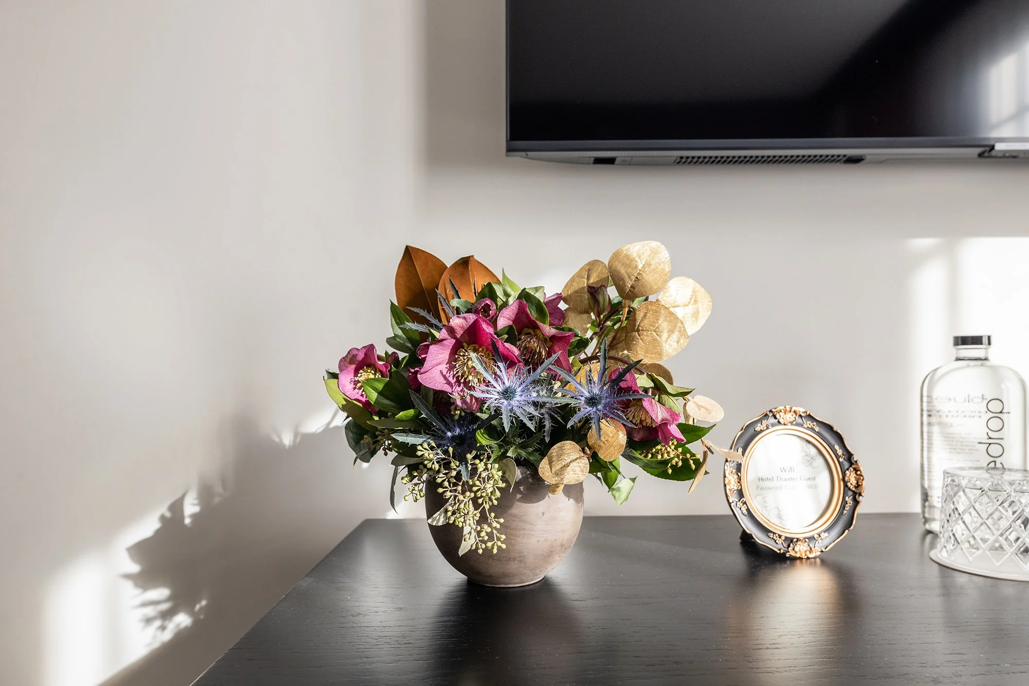 A flower arrangement in a round vase on a black table, with a decorative picture frame, a bottle of water, and a glass on the right, and a TV mounted on the wall above.