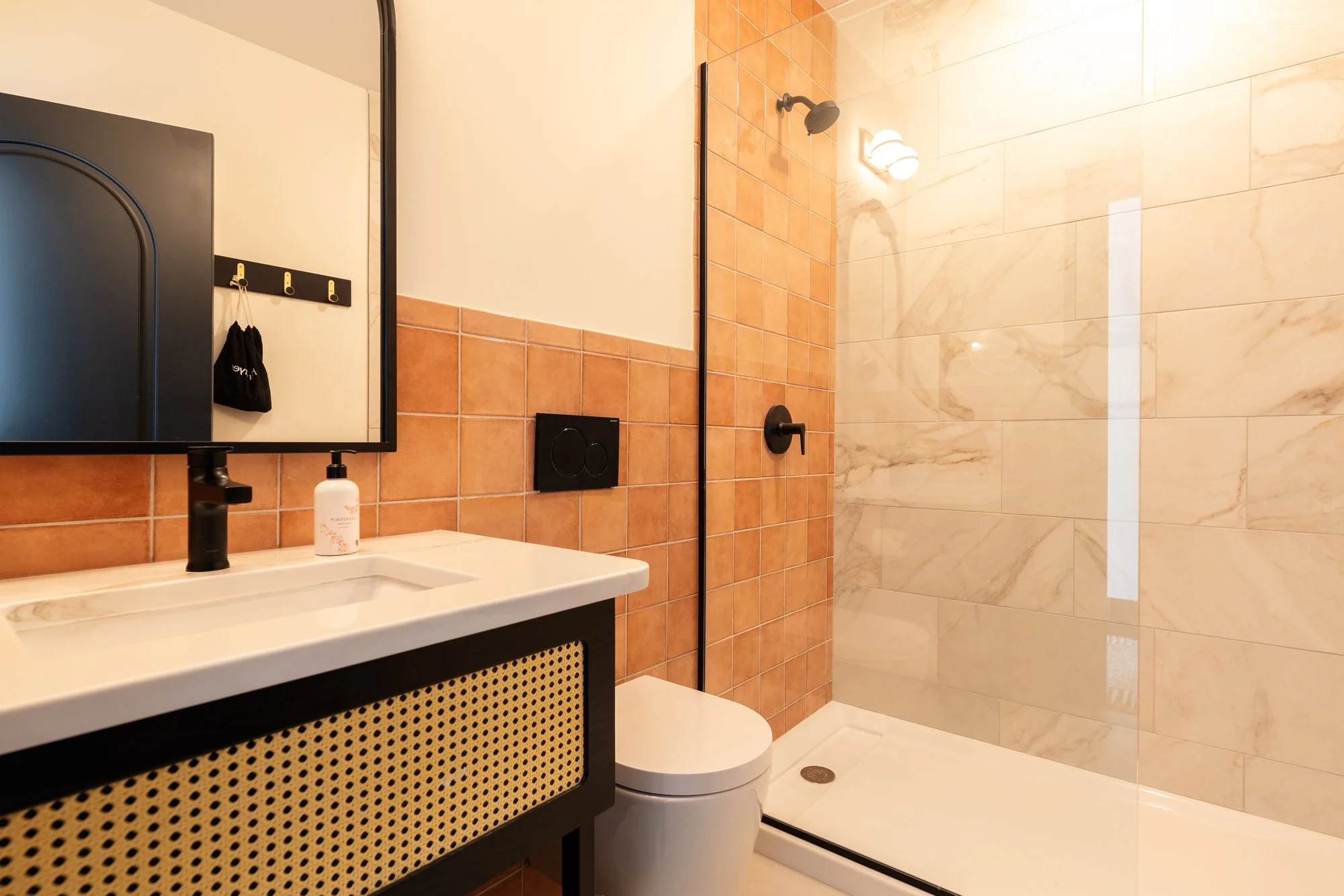 Modern bathroom with a black-rimmed mirror, white sink, black faucet, liquid soap dispenser, orange tiles behind the sink, a white toilet, and a walk-in shower with beige marble tiles, a black showerhead, and a black shower handle.