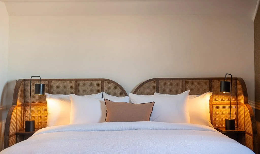 Bedroom with rattan king headboard, black end table lamps, white linens, and a blush accent pillow in the Downtown Luxury Condo at Hotel Thaxter in Portsmouth, New Hampshire.