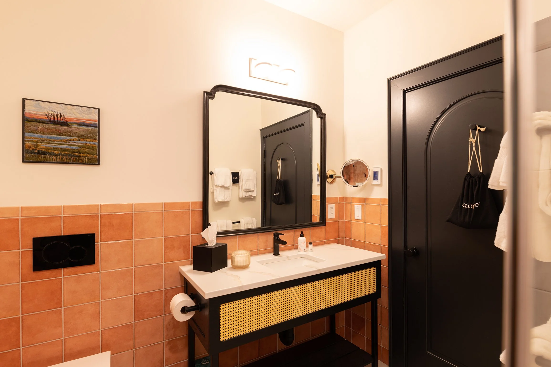A modern bathroom with a peach-colored tile wall, a black framed mirror above a white vanity with black accents, a faucet, and various toiletries. A black door has a bag hanging on it, and there are white towels and artwork on the walls.
