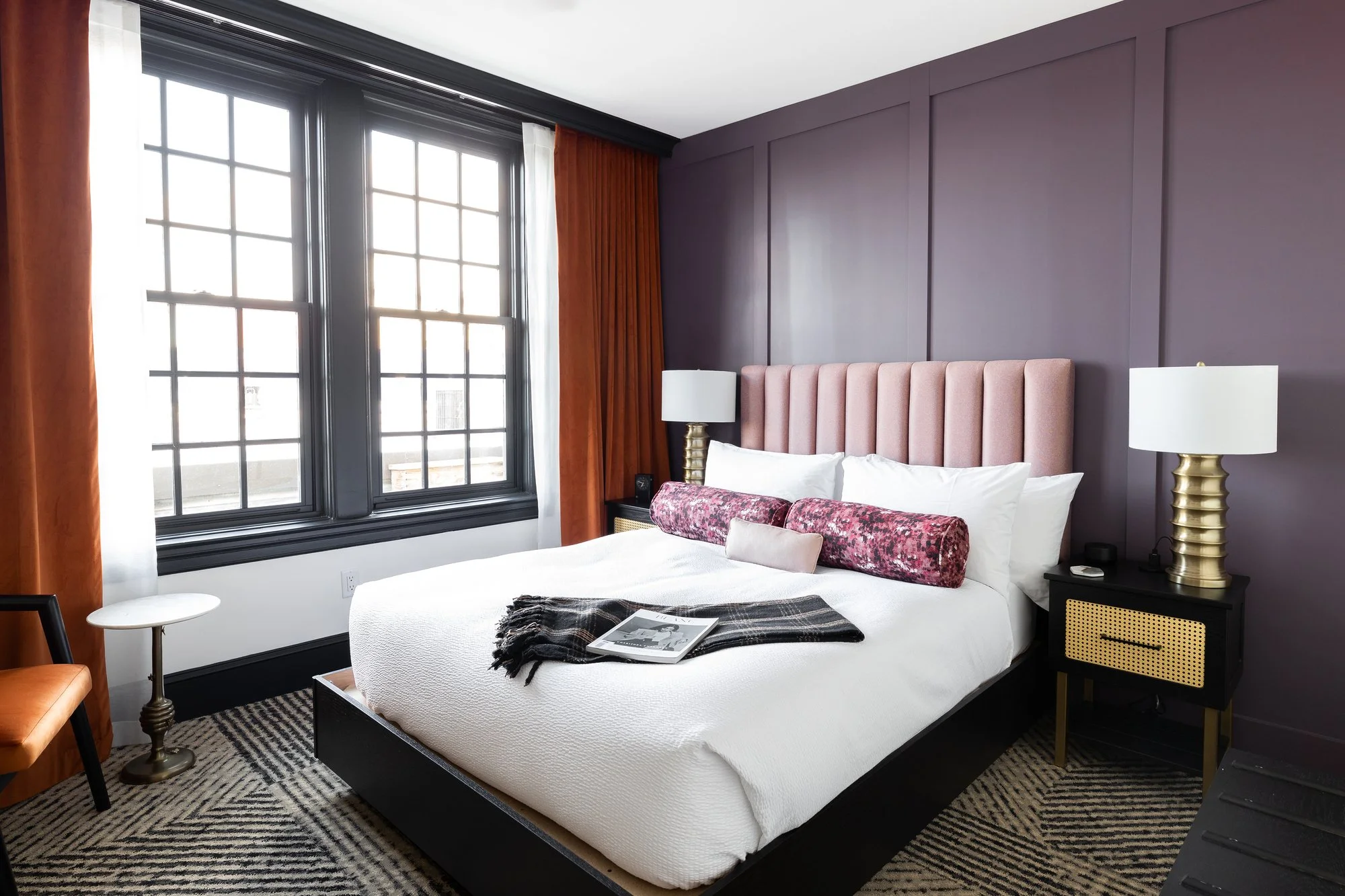 Modern bedroom with a large bed featuring a pink upholstered headboard, white bedding, floral and beige cushions, black nightstands with gold lamps, large black-framed windows, orange curtains, a black chair, white side table, and a zebra-patterned r