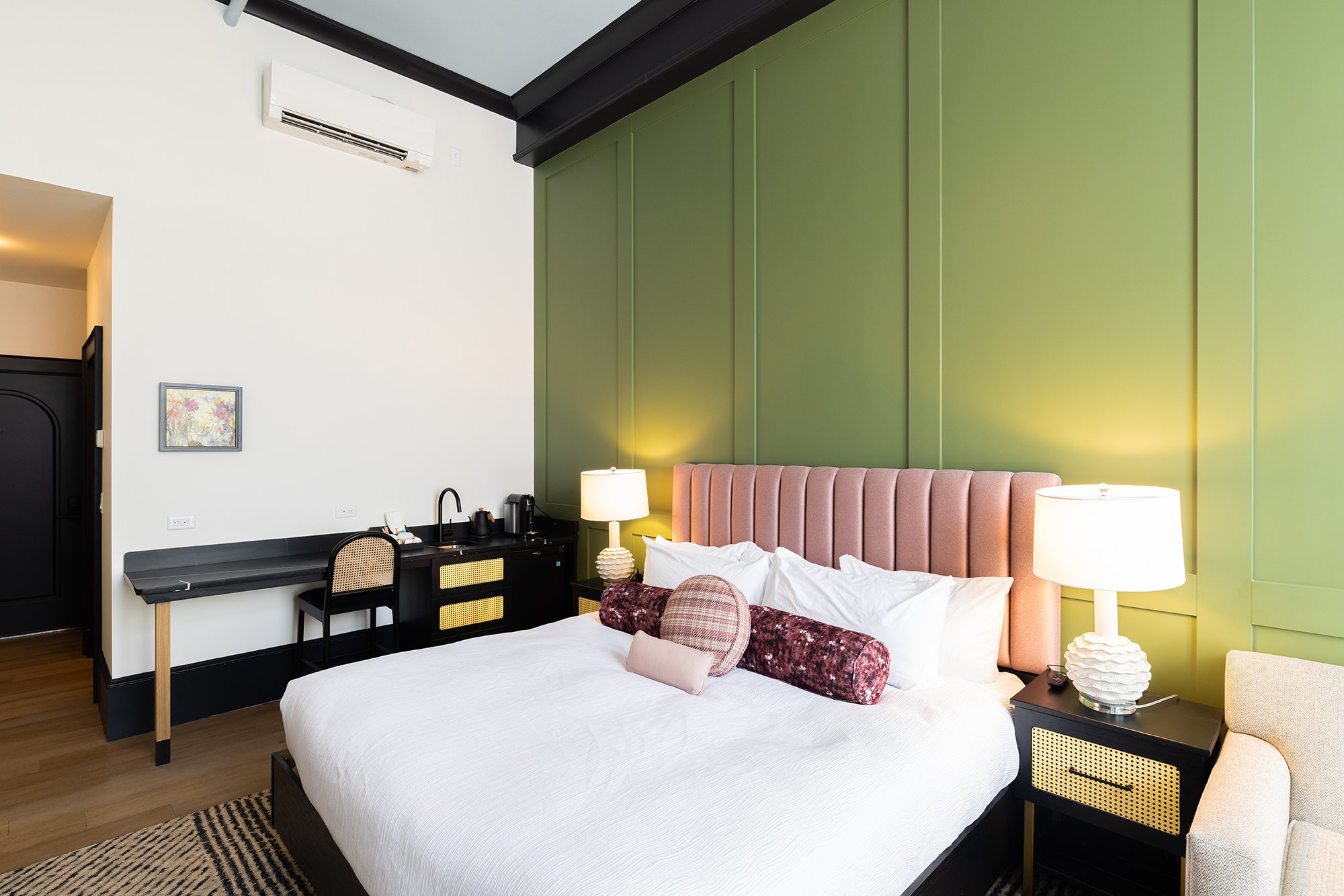 Interior of a modern bedroom featuring a bed with a pink upholstered headboard, two white lamps, a nightstand, and a green accent wall.