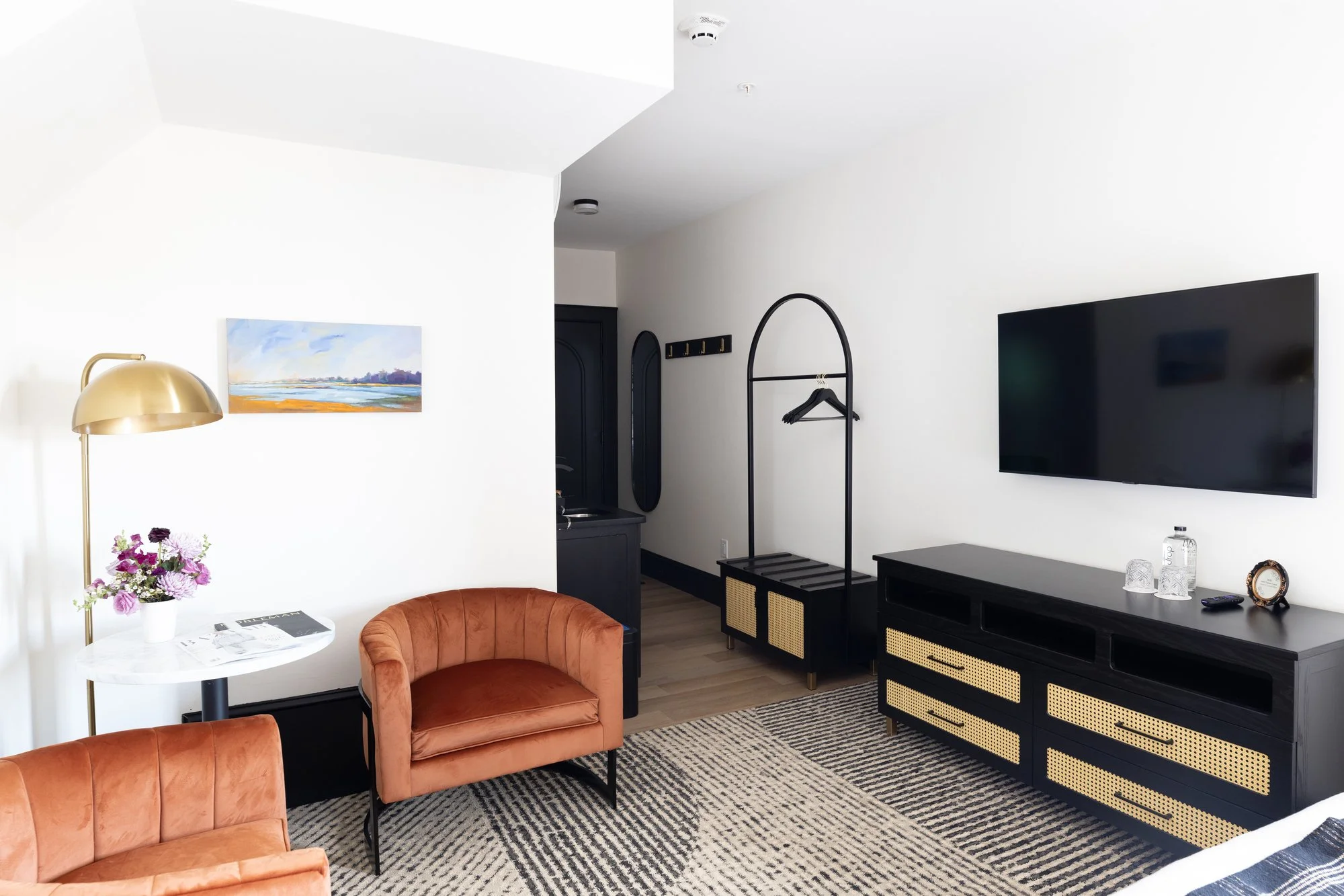 Modern living room with white walls, two pink velvet armchairs, a white side table with a flower arrangement, a gold lamp, a large flat-screen TV on a black TV stand, a black coat rack, and a black cabinet. There is a abstract landscape painting on t