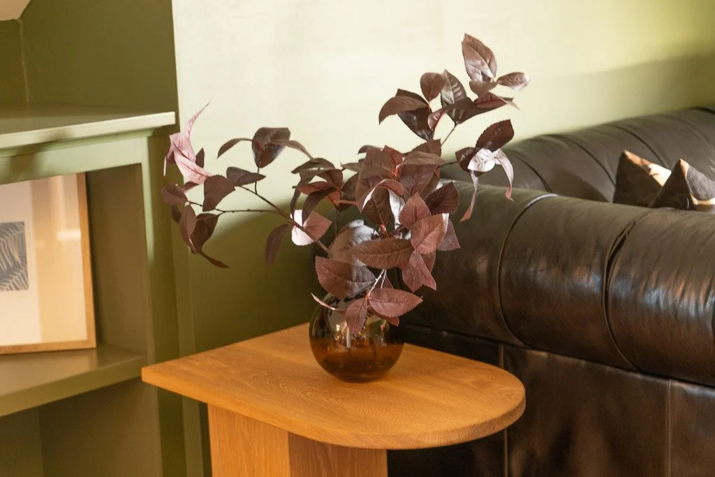 End table with decorative purple foliage near black tufted couch and green accent wall in the Downtown Luxury Condo at Hotel Thaxter in Portsmouth, New Hampshire.