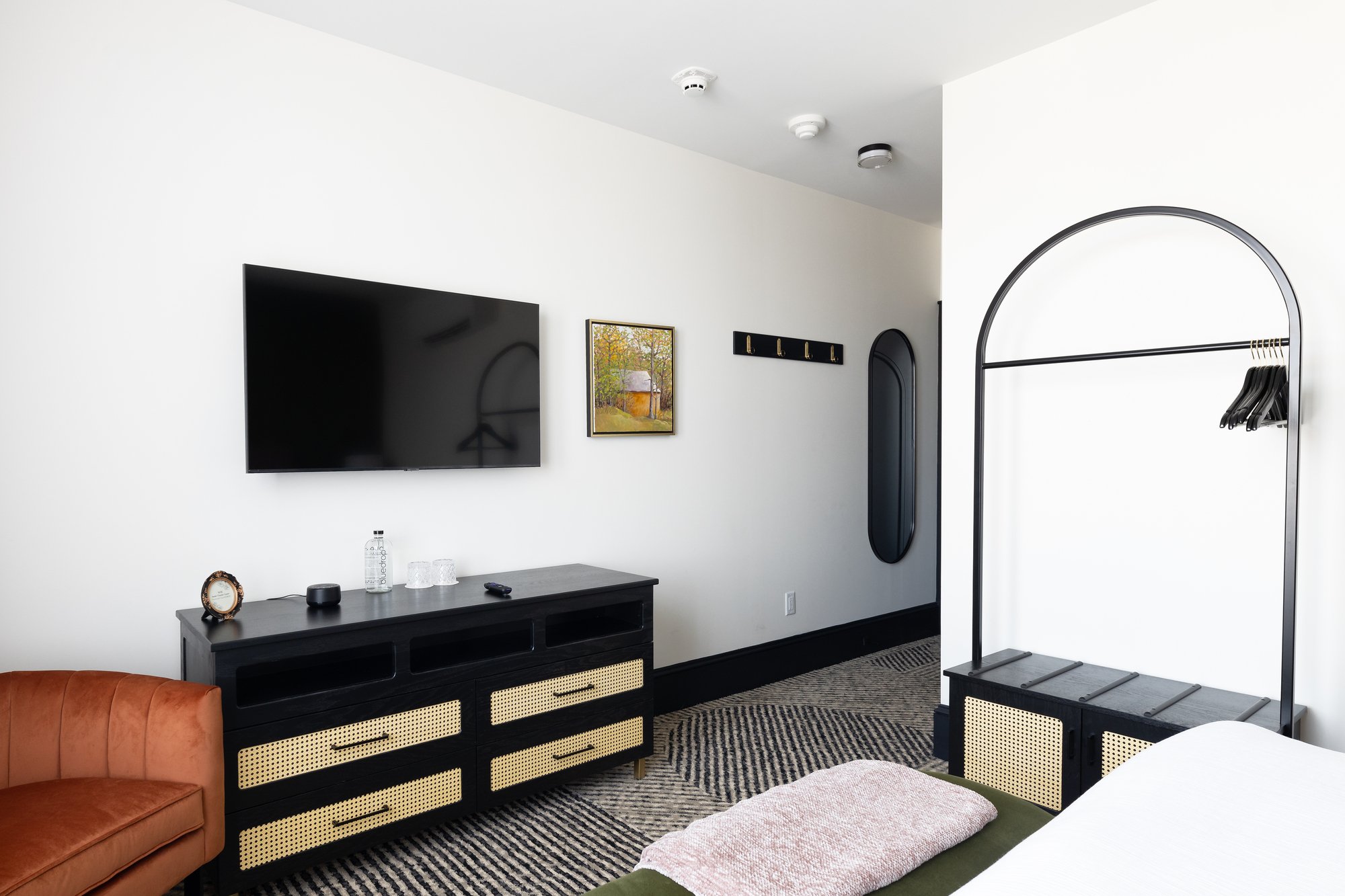 Interior of a modern bedroom with a wall-mounted TV, framed artwork, a black bench, a coat rack, and a clothing rack with hangers. A part of a bed with a pink blanket and an orange chair are also visible.